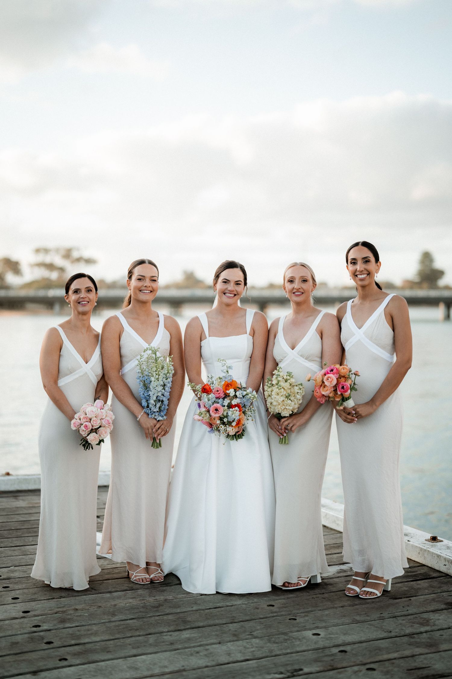 Bridal party in white dresses poses with bouquets on wooden pier by waterfront.