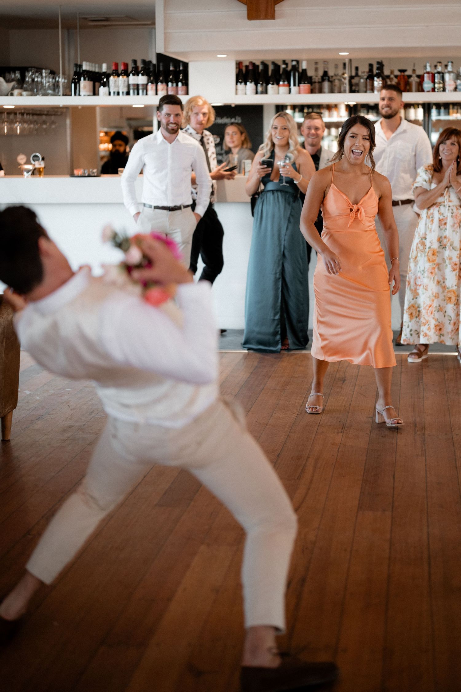 Wedding guests dancing and celebrating on a wooden dance floor with a bar in the background.