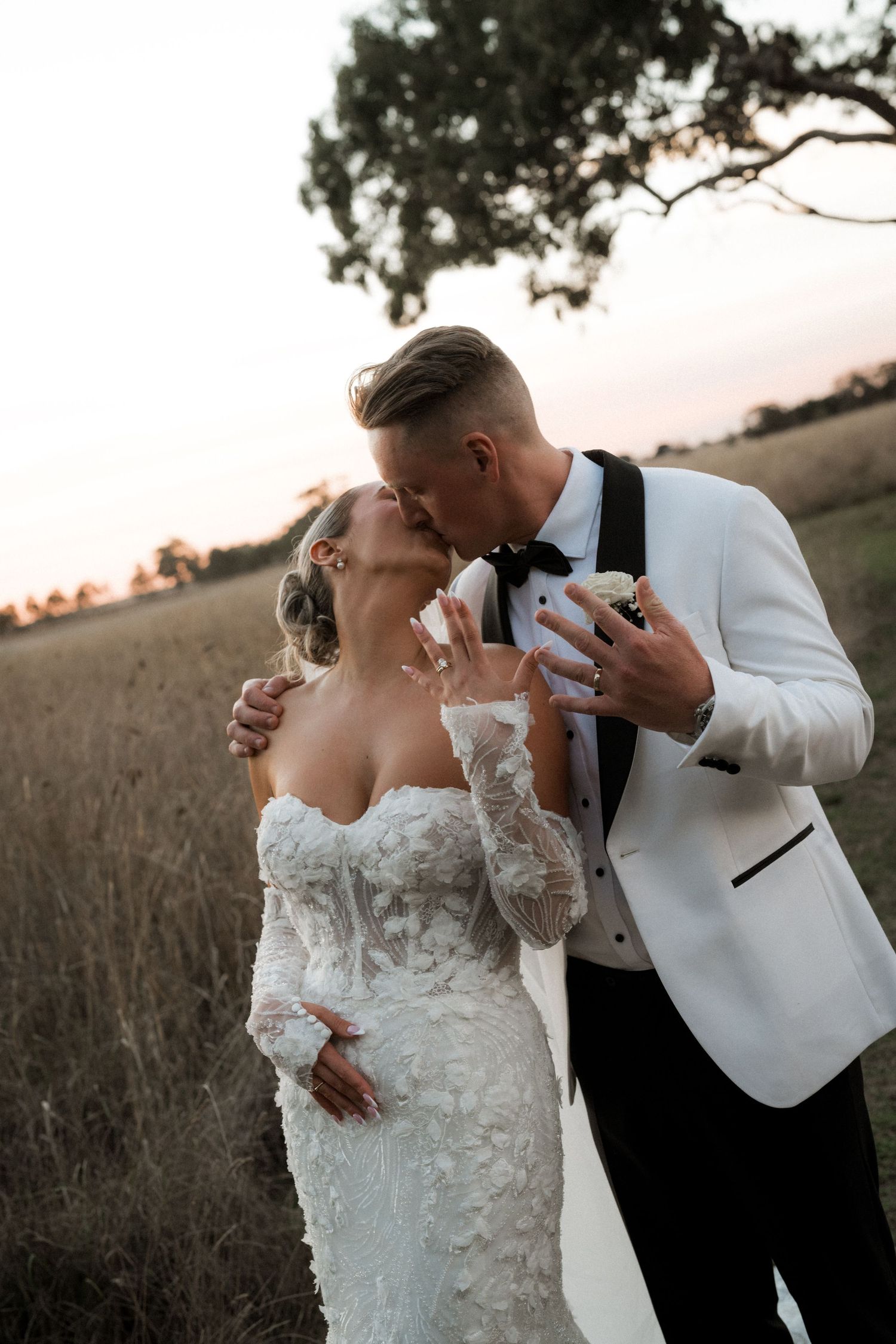 A romantic sunset portrait of newlyweds embracing in a field, with the groom wearing a white dinner jacket.