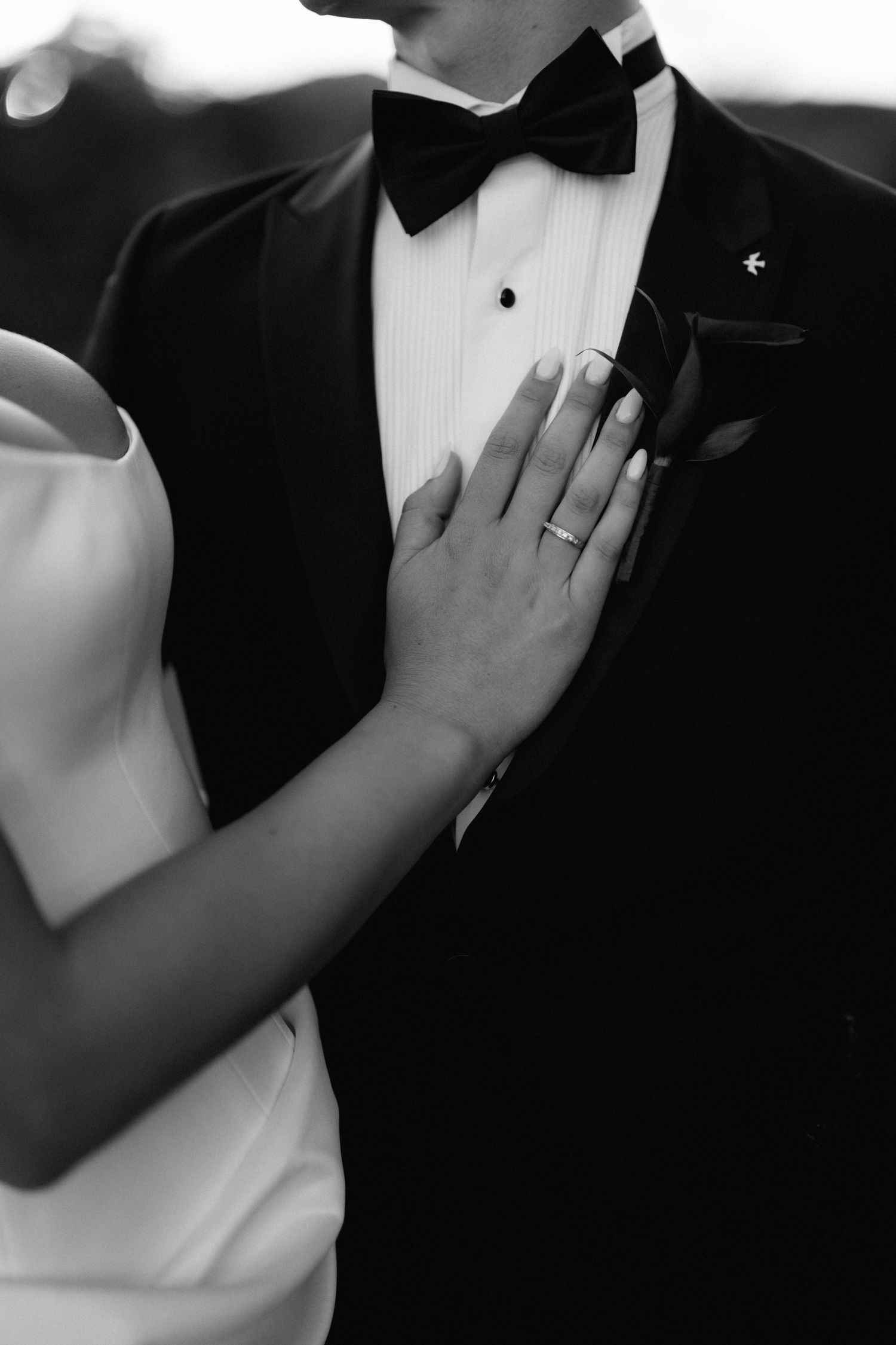Close-up of a delicate hand resting on a tuxedo-clad chest showing a wedding ring.