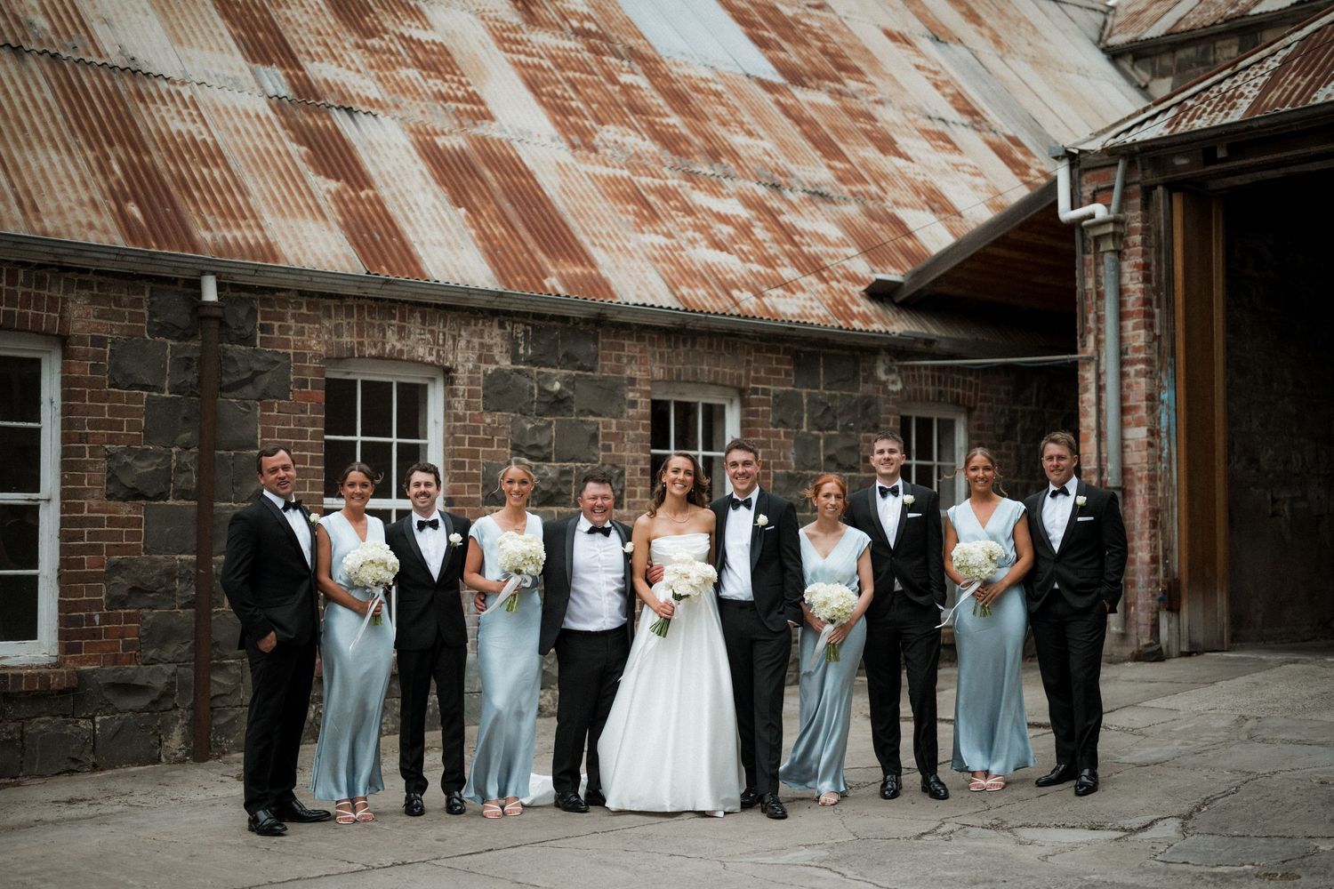 Wedding party poses together against rustic brick building with weathered metal roof.