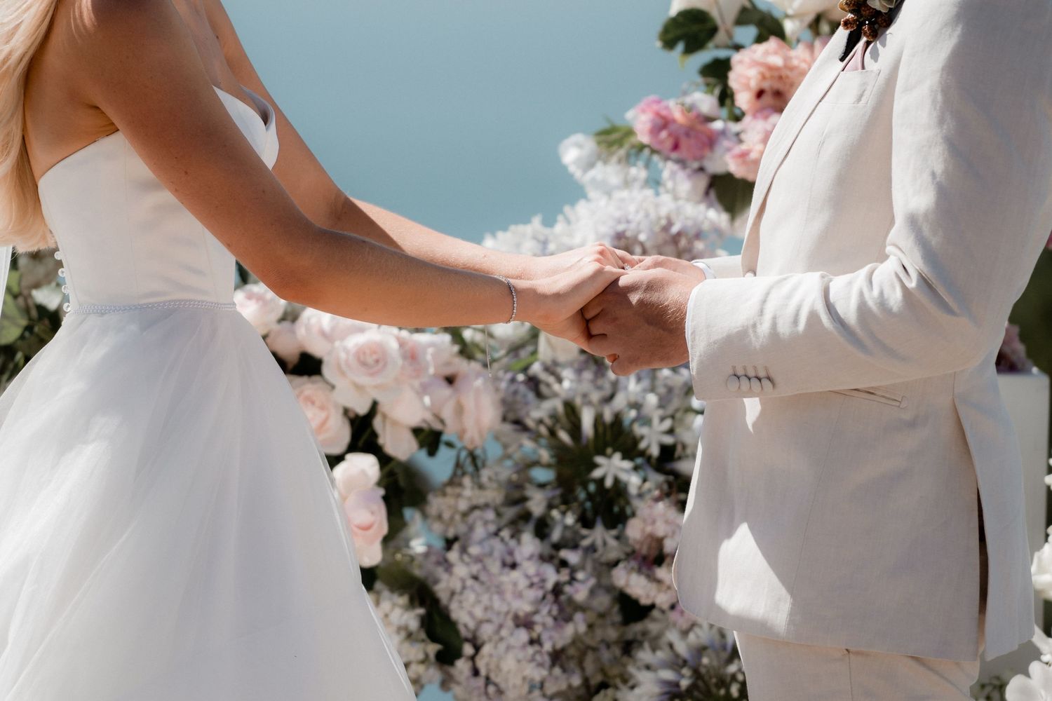 A couple holds hands during their wedding ceremony in front of a pink and white floral arch backdrop.