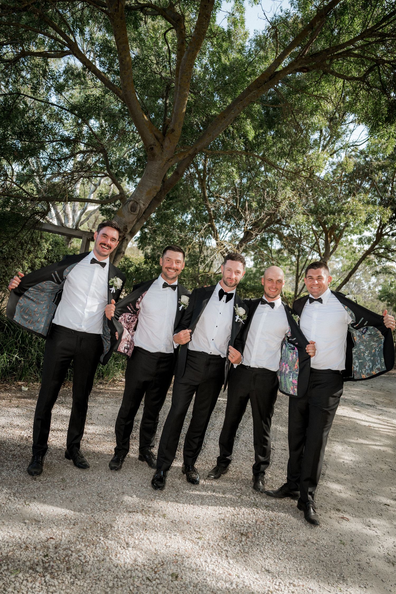 A group of groomsmen in formal black and white attire pose together under trees in an outdoor setting.