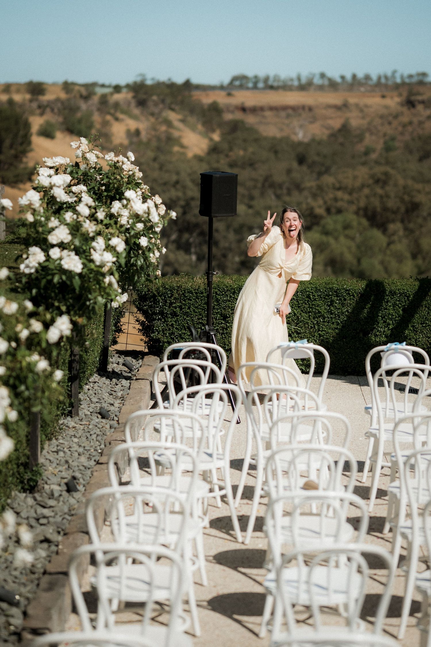 Woman in cream dress poses playfully among rows of white chairs at an outdoor garden wedding ceremony.