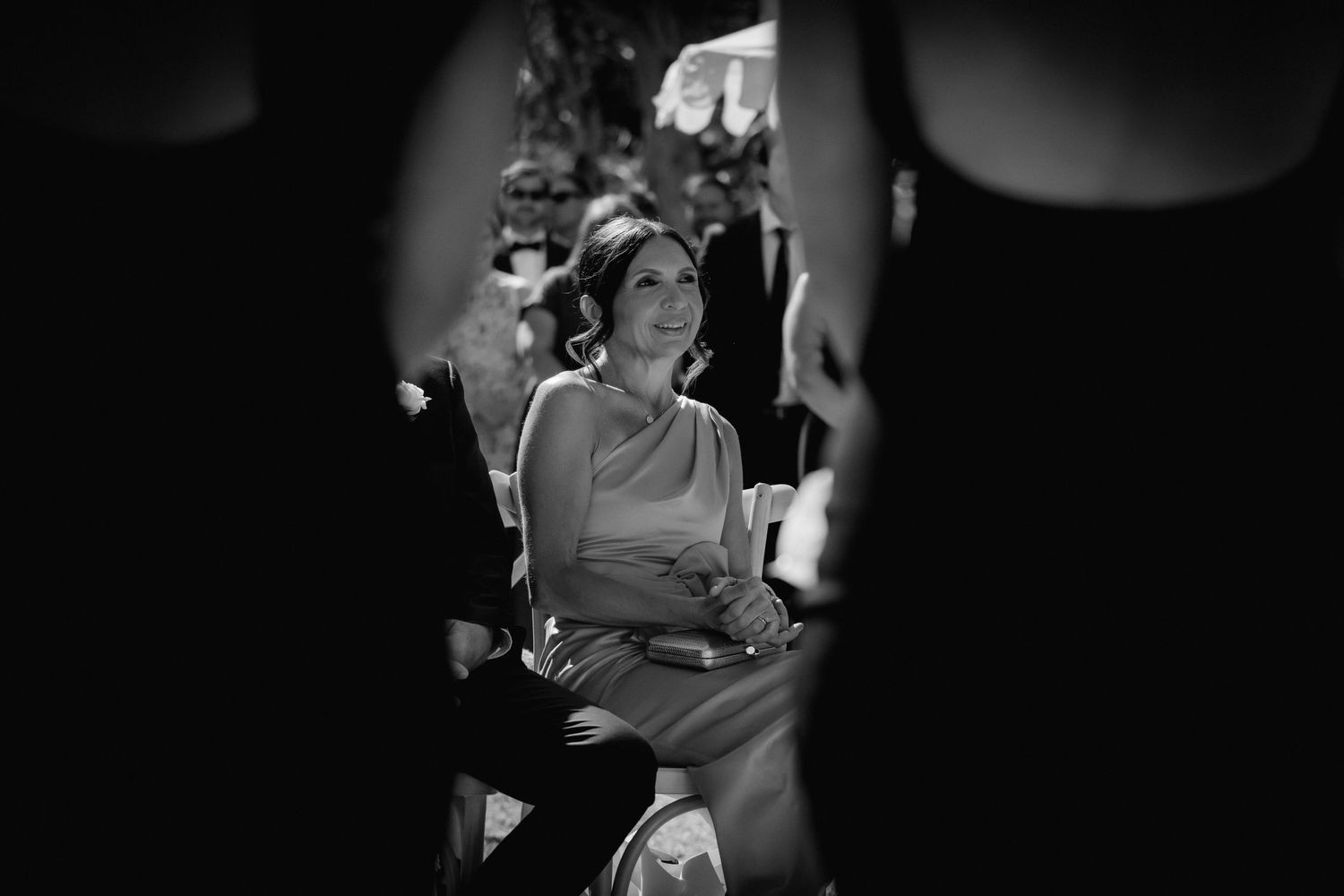 Black and white photo of a woman smiling while seated at an outdoor wedding ceremony.