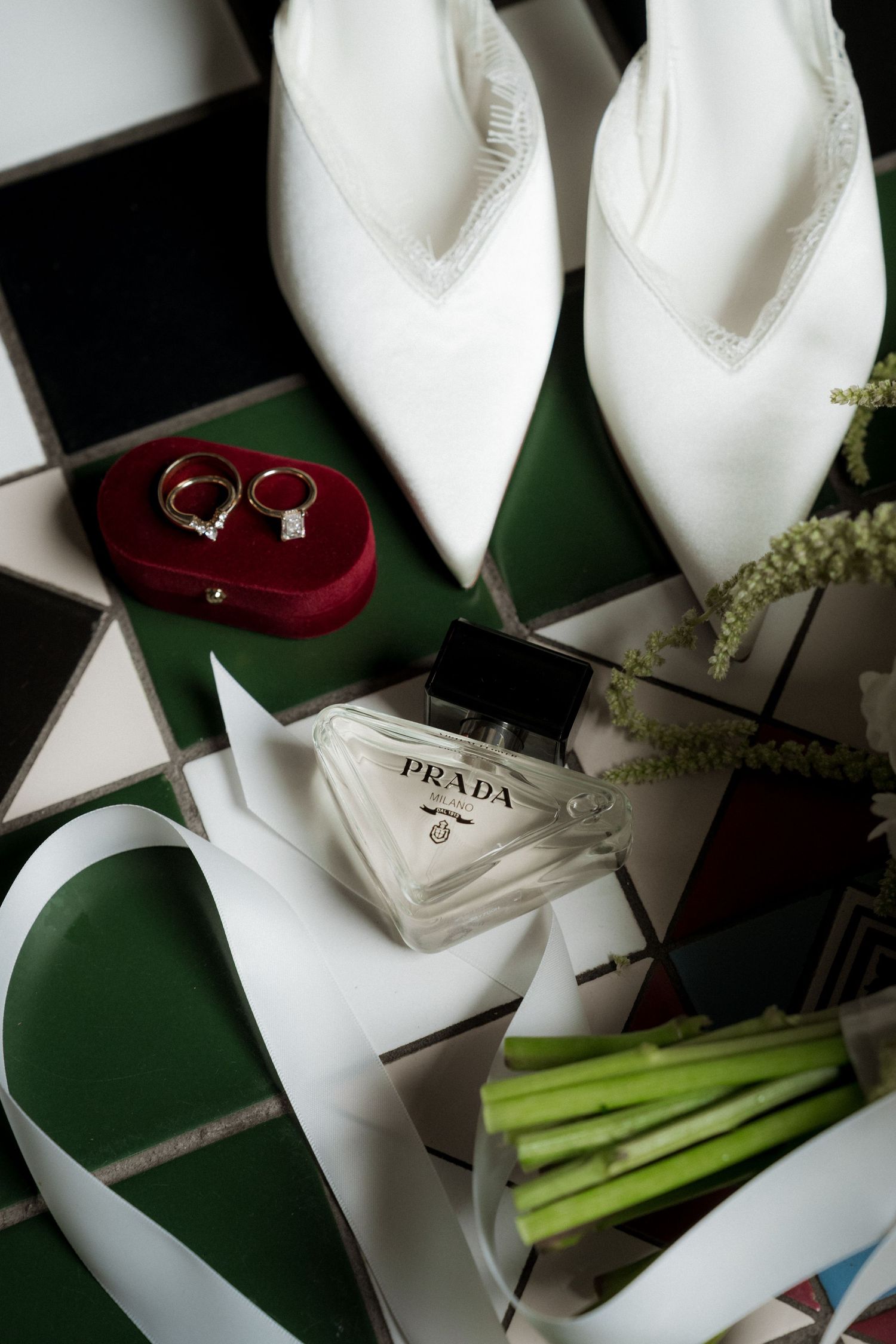 White Prada bridal shoes, gold wedding rings in red velvet box, and Prada perfume arranged on green geometric tiles.