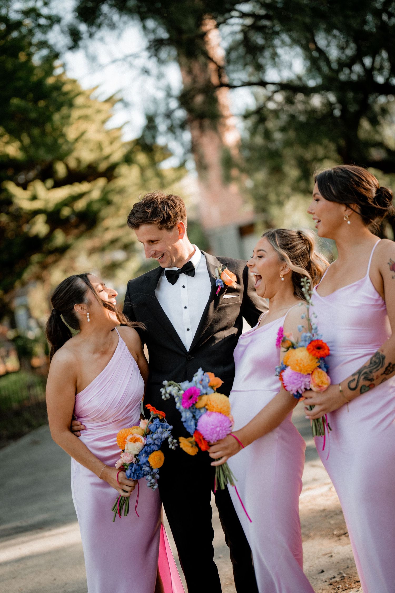 Bridal party wearing lavender dresses hold vibrant rainbow bouquets while sharing laughs outdoors.