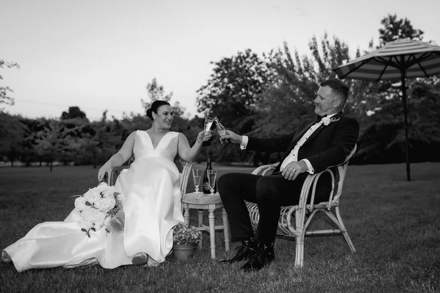 A black and white photo of newlyweds relaxing on outdoor furniture in a garden setting.