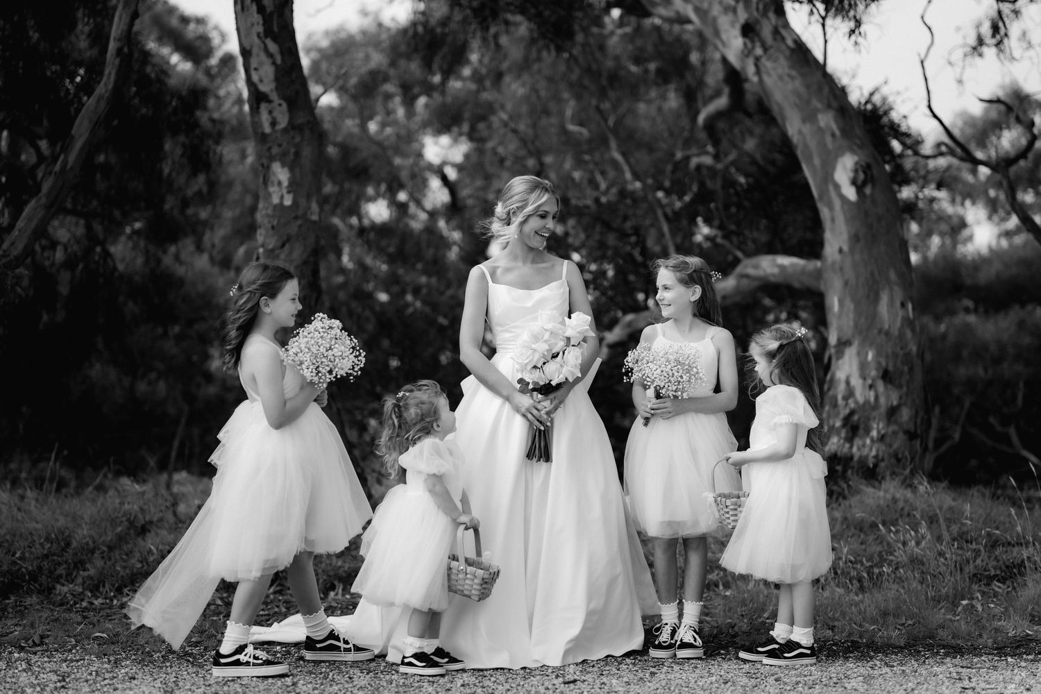 Black and white wedding photo showing a bride and little flower girls in white dresses under trees.