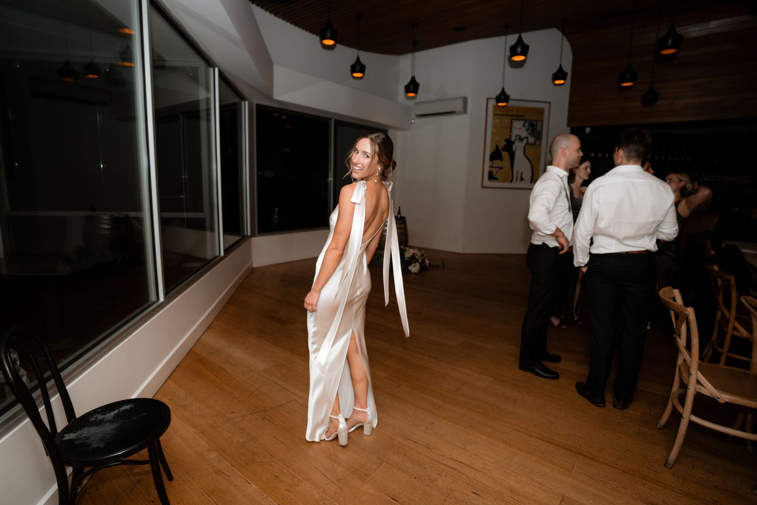 Wedding guest in flowing white dress standing on wooden dance floor with others in background.
