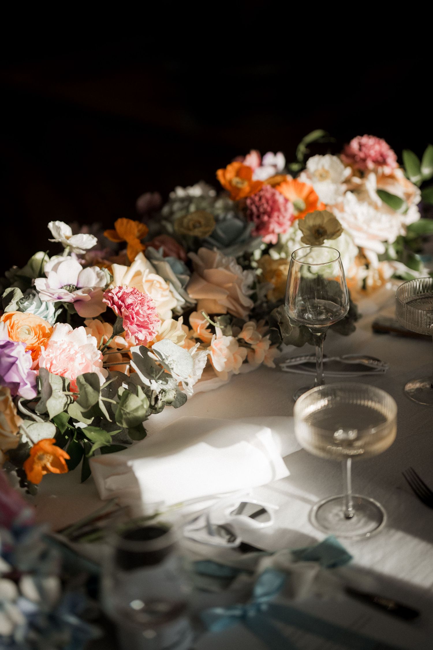 Elegant wedding table with colorful floral centerpiece and champagne coupe glasses in dramatic lighting.