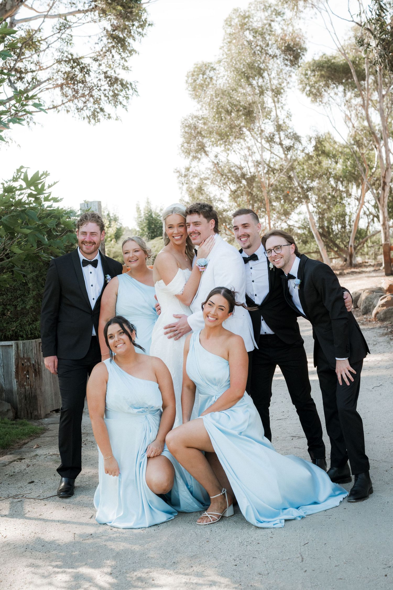 Wedding party group photo with bride, groom, bridesmaids in blue and groomsmen in black tuxedos outdoors.