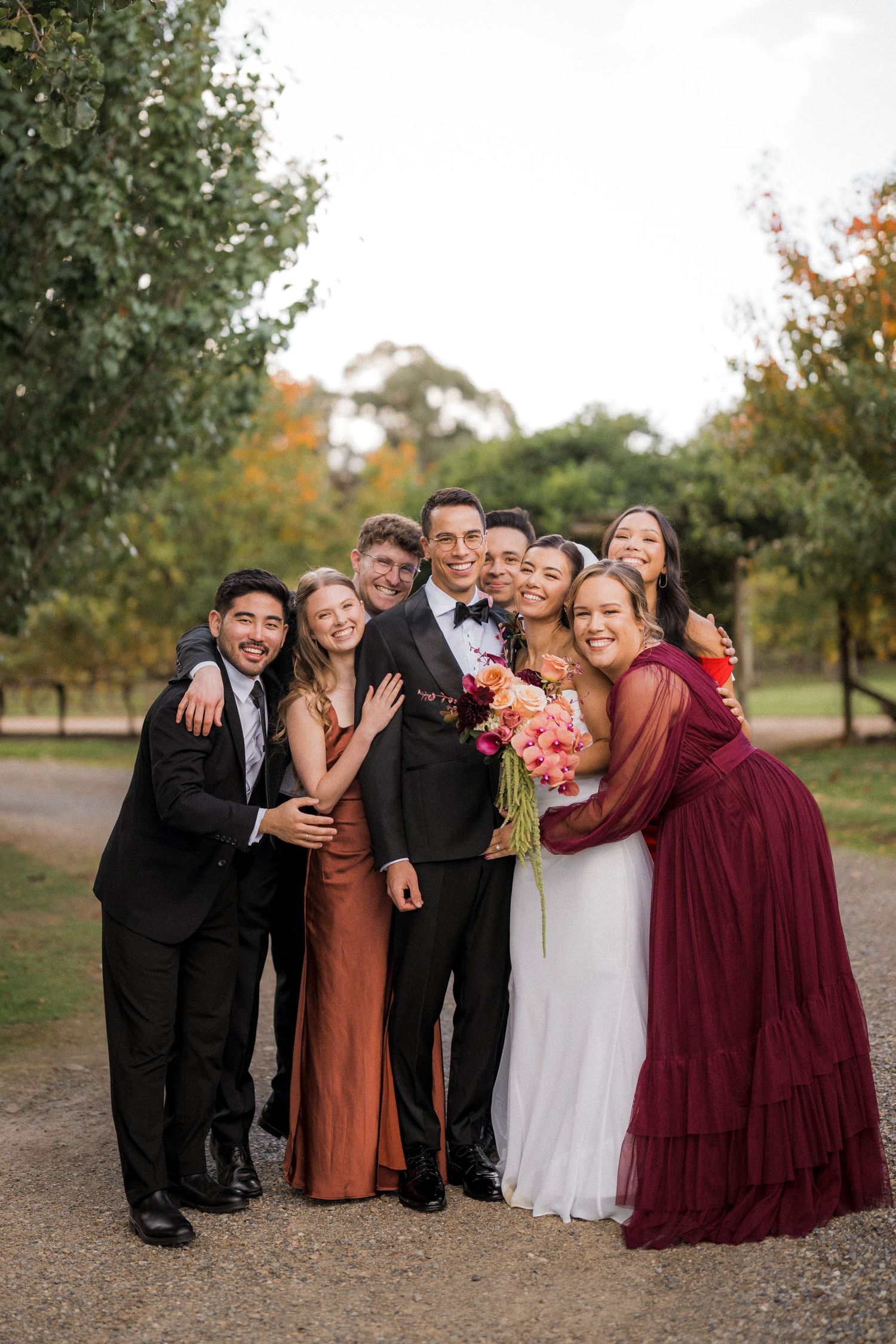 Bride and groom surrounded by smiling wedding party outdoors among autumn trees.