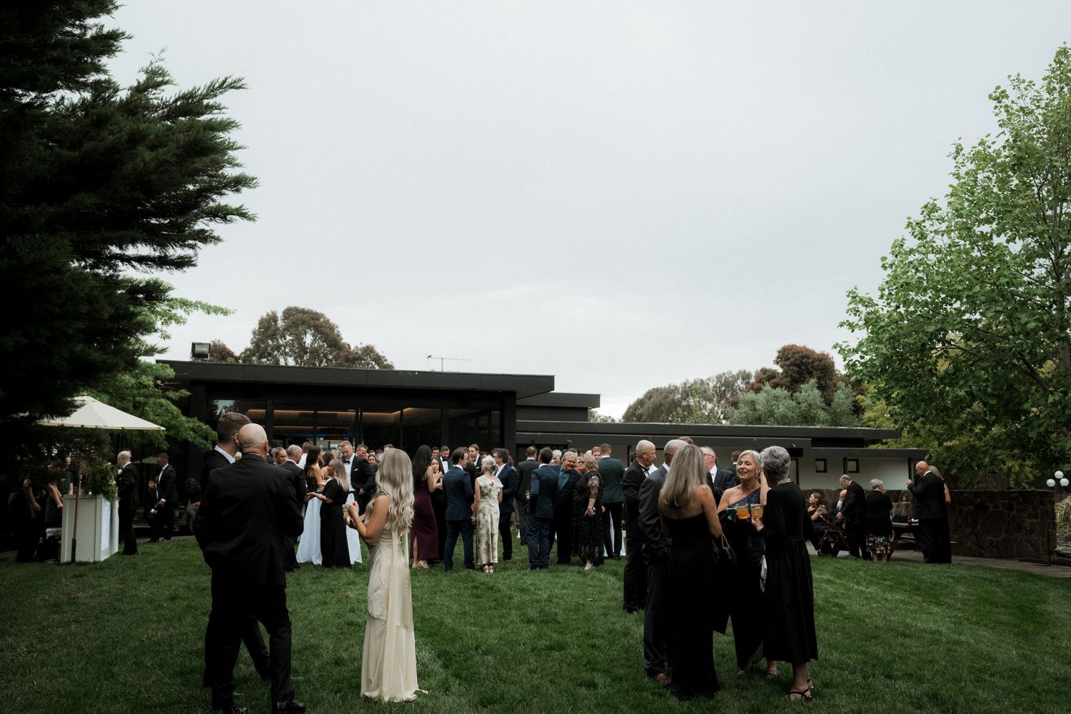 Outdoor wedding reception with guests gathered on a lawn near modern architecture and trees.
