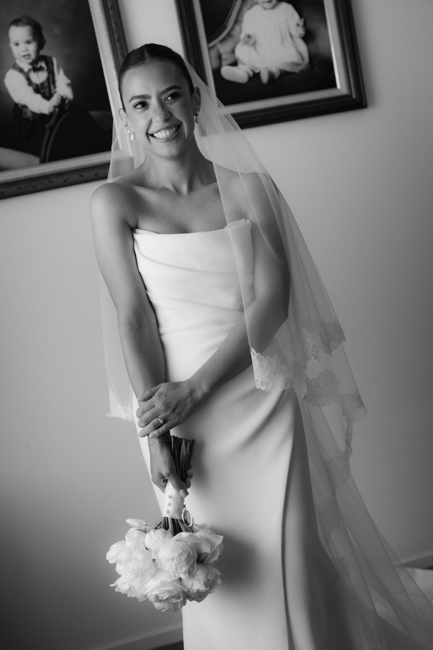 A bride in an elegant white wedding dress holds a bouquet while standing in black and white photography.