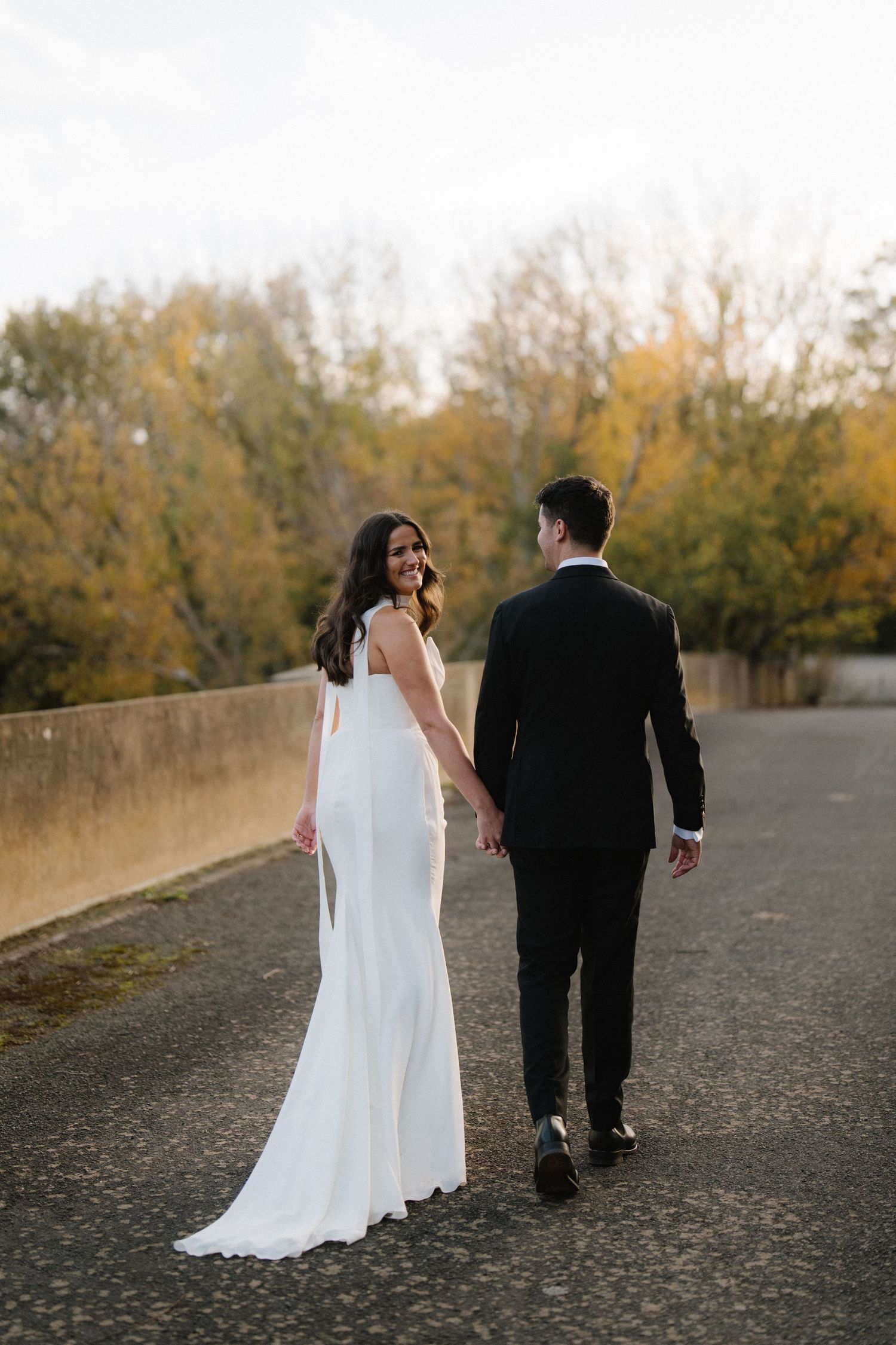 Bride and groom walking together on a path surrounded by autumn trees at sunset.