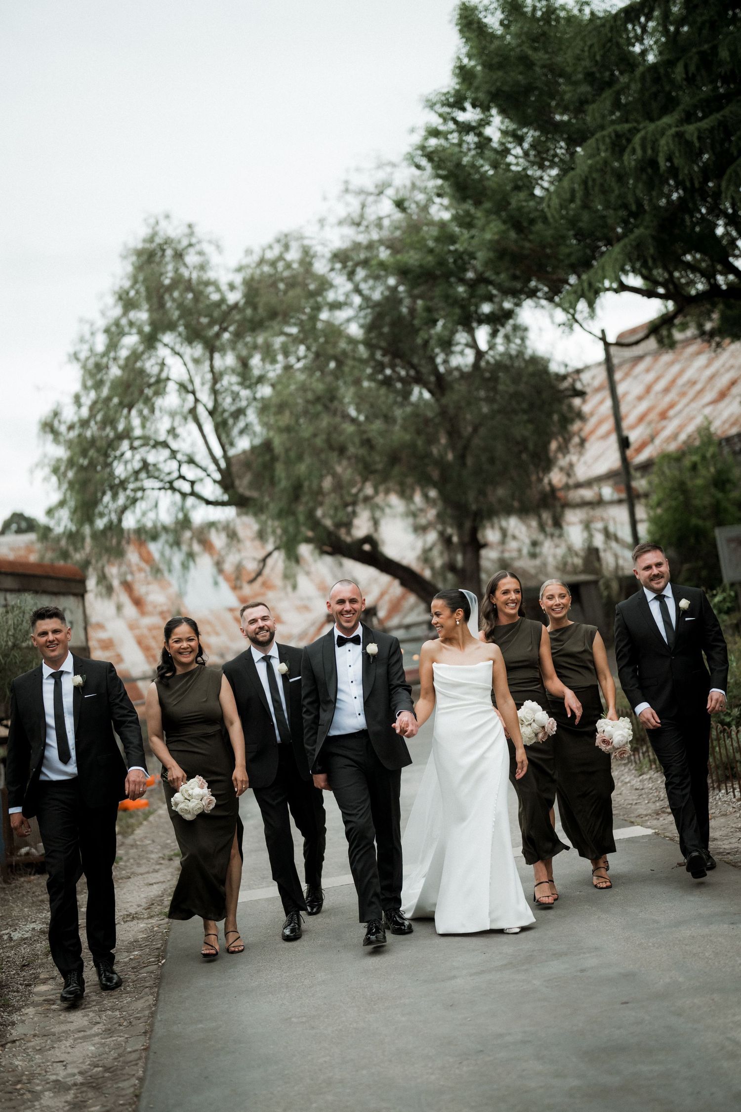 Wedding party walking together on a path with trees and buildings in the background.