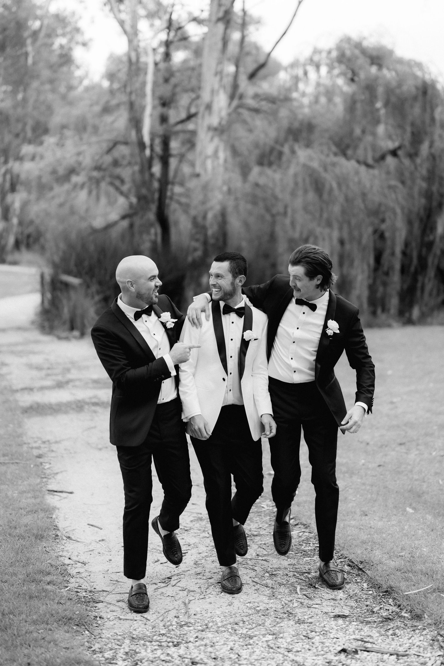 Groomsmen in black tuxedos performing synchronized leg kicks outdoors in black and white photography.