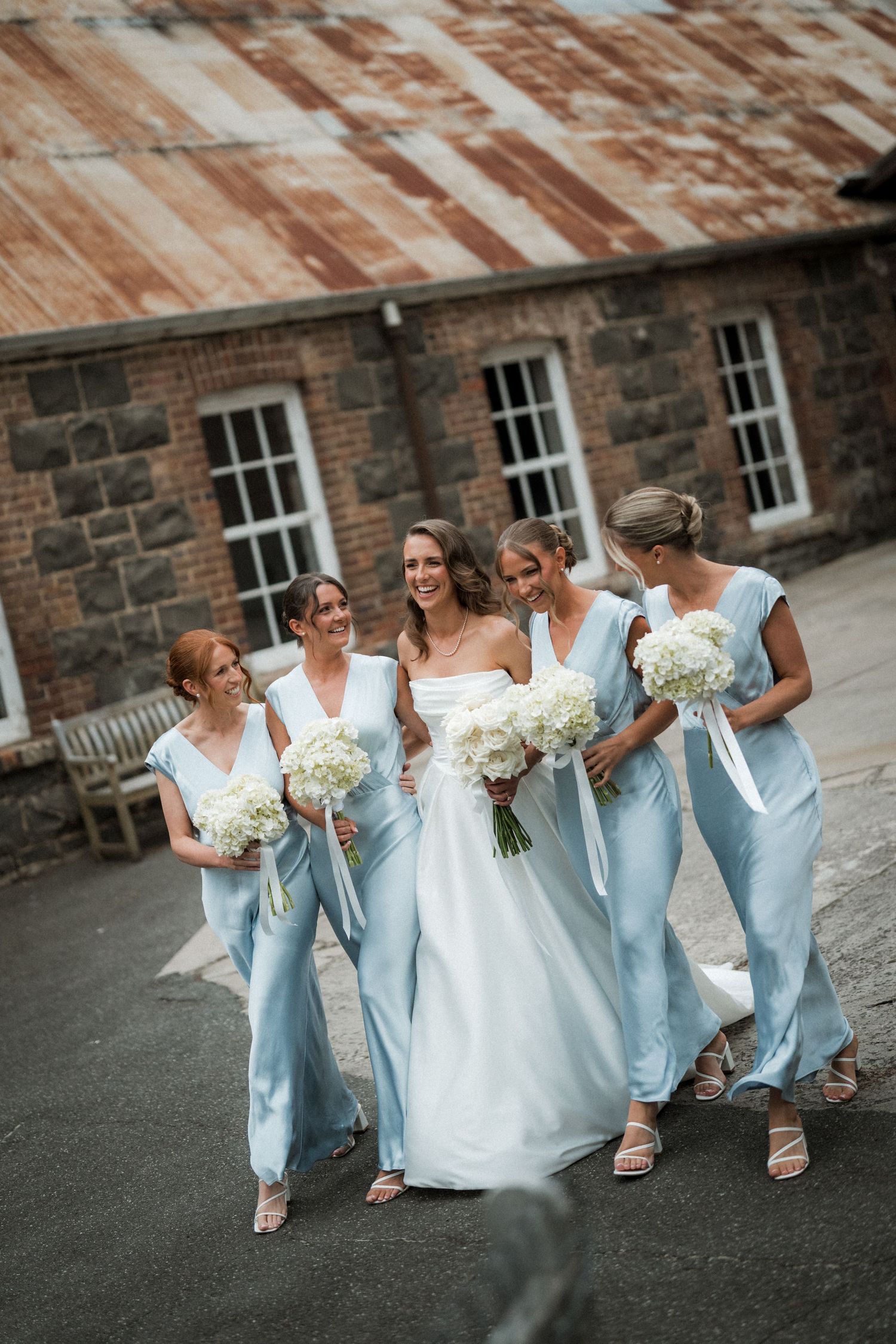 Bridesmaids in light blue dresses walk together holding bouquets against historic stone building.