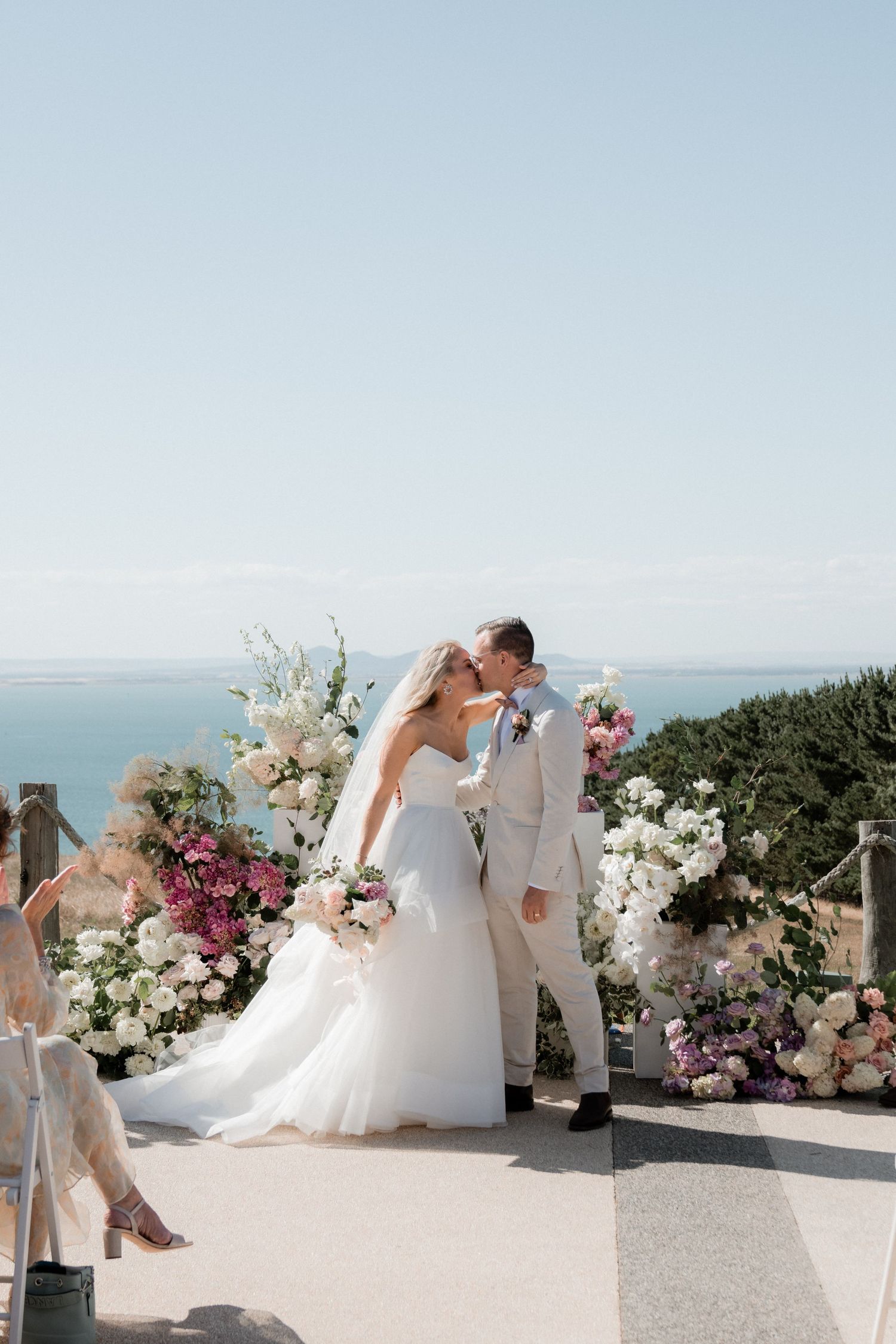 A romantic wedding ceremony overlooking the ocean with elegant white and pink floral arrangements on a sunny day.