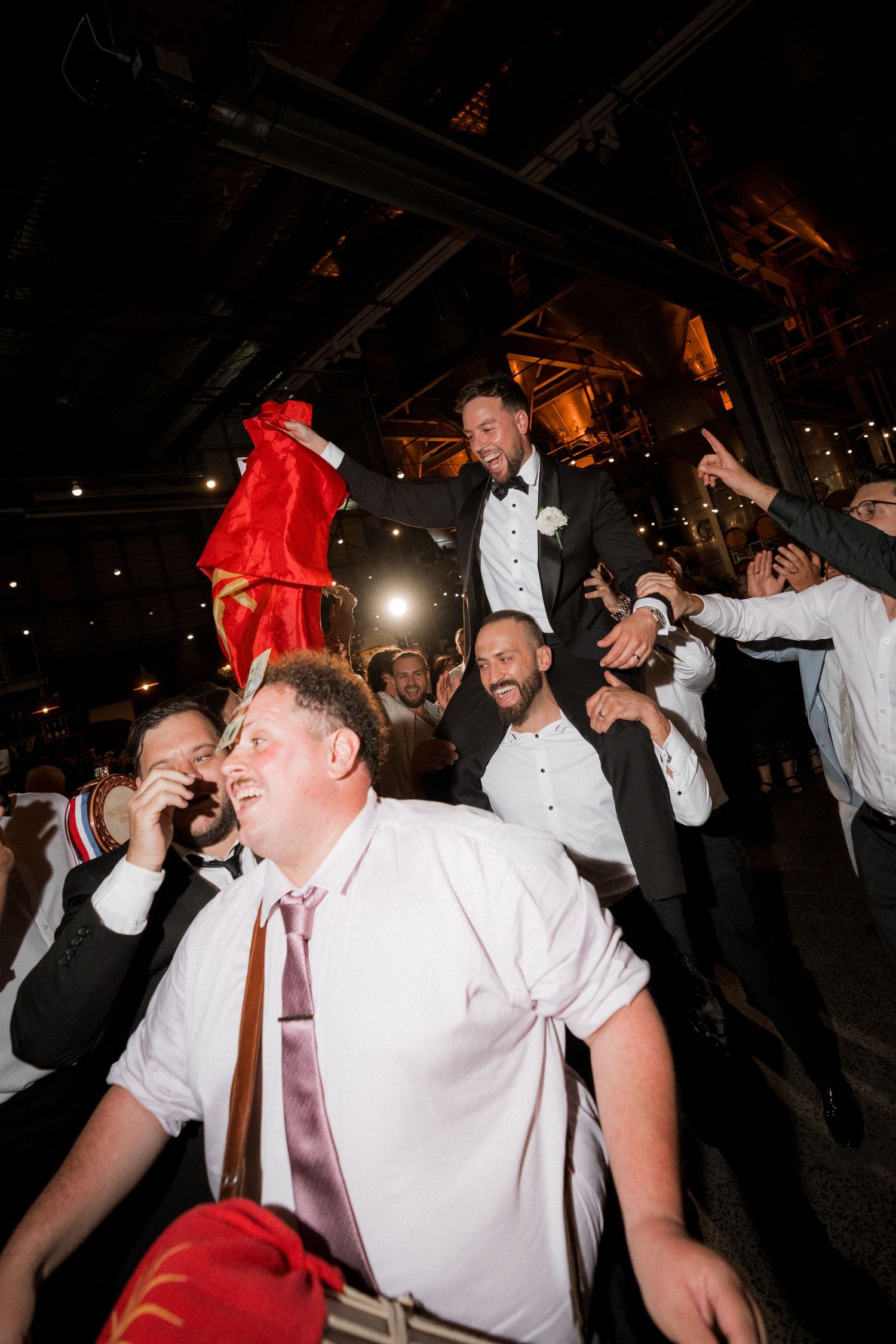 Wedding guests in white shirts dance energetically on a dark dance floor at a reception celebration.