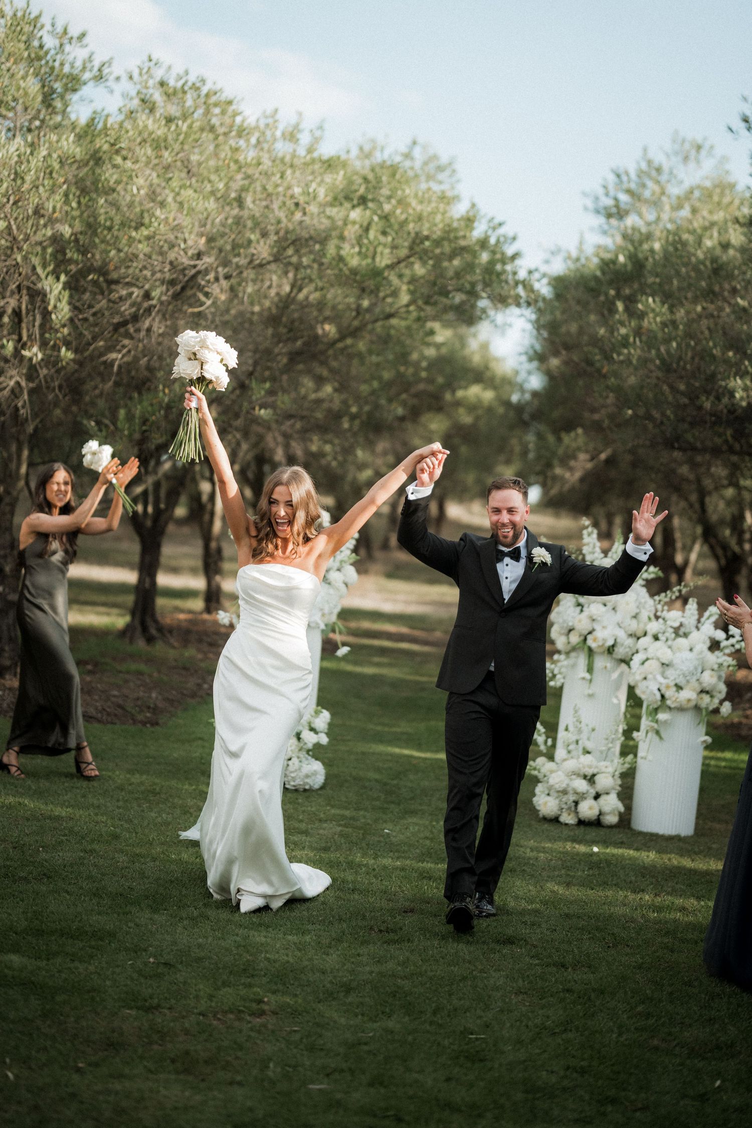 Newlyweds celebrate with raised arms while walking through an olive grove after their ceremony.