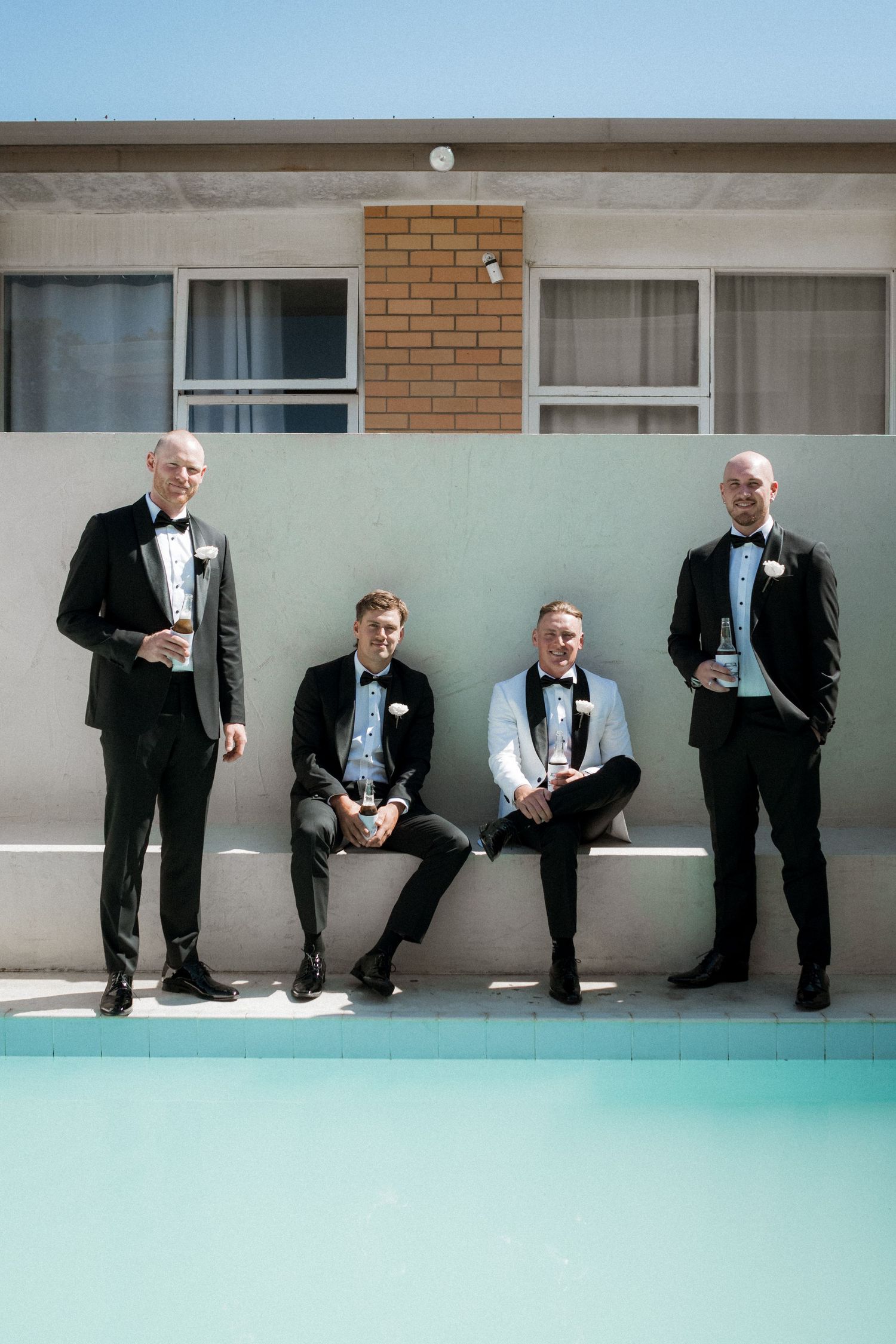 Groomsmen in tuxedos sitting casually along a poolside wall in a fun group photo.