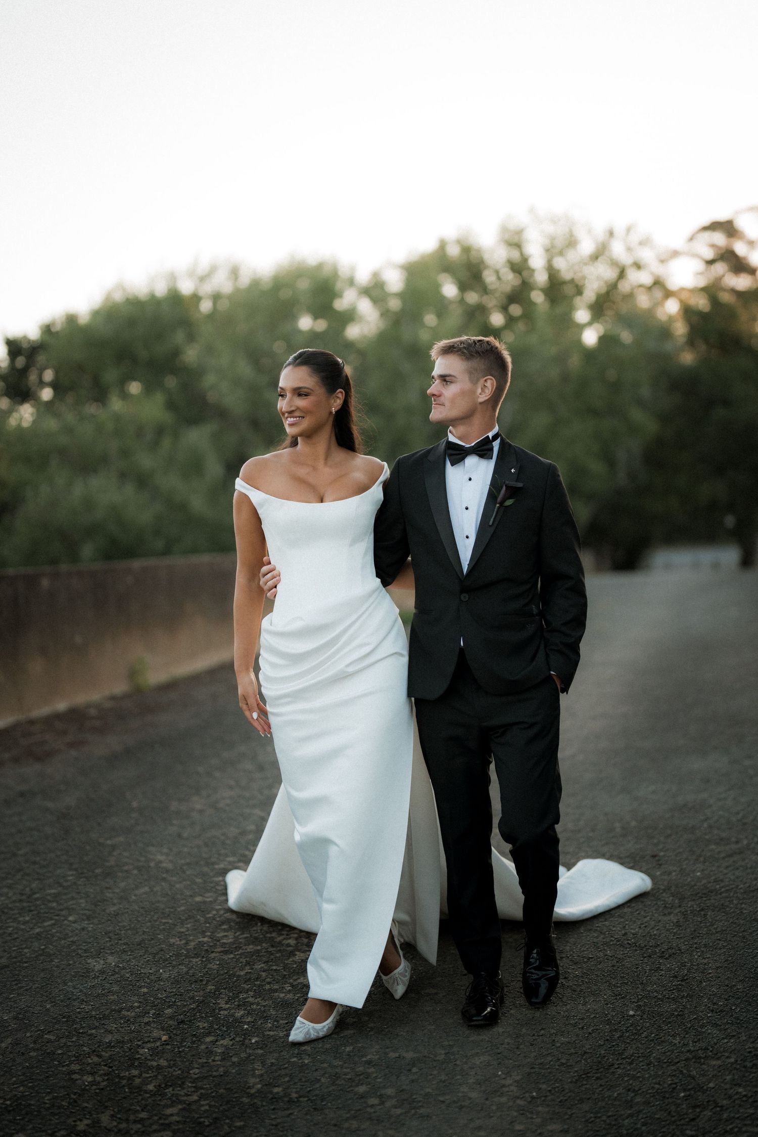 Bride in white dress and groom in tuxedo walking together on path.