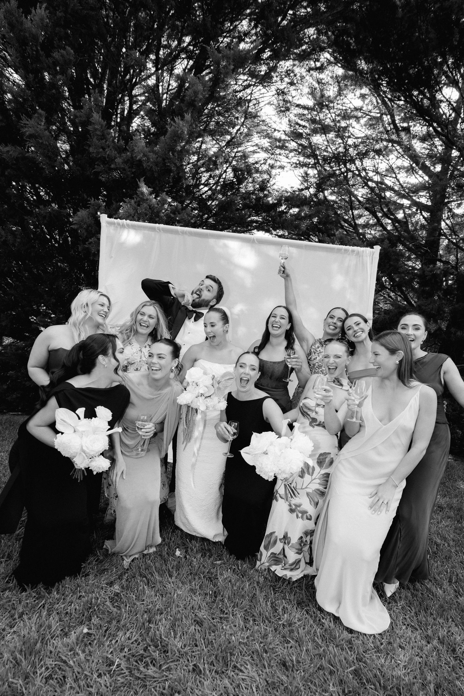 Black and white group photo of bride, groom, and bridesmaids celebrating joyfully outdoors.