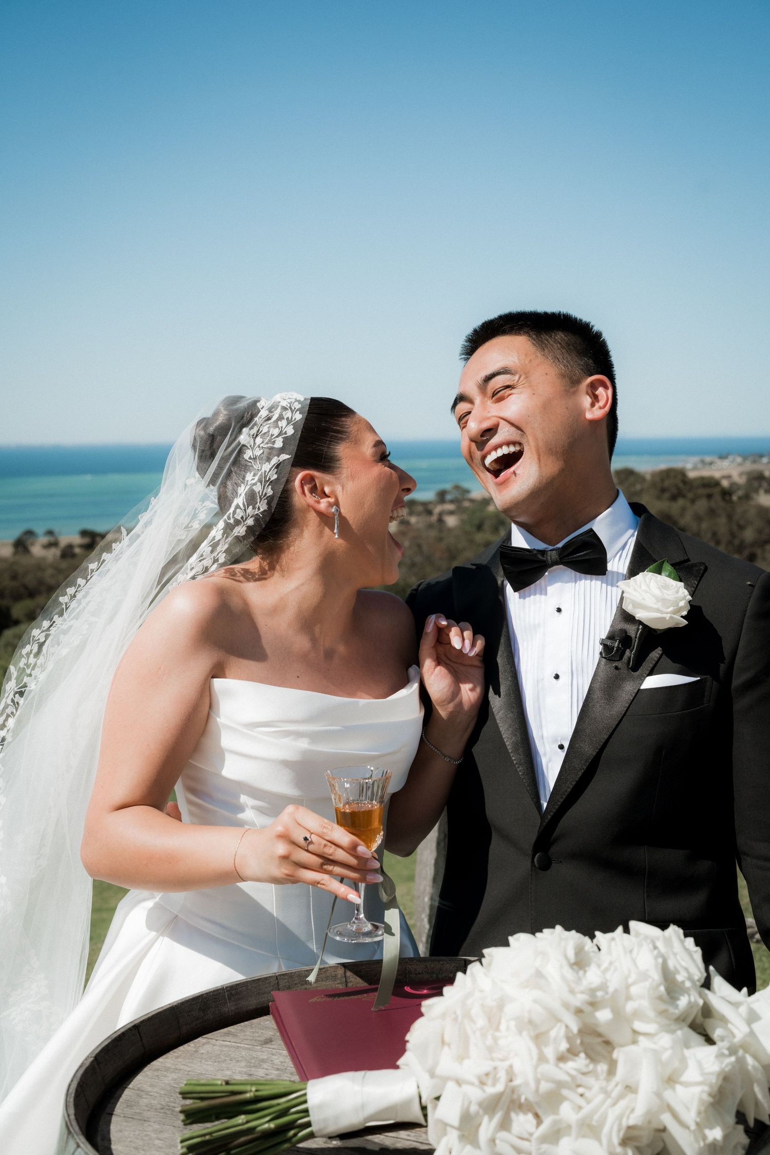 Joyful wedding couple sharing drinks and laughter against an ocean backdrop.