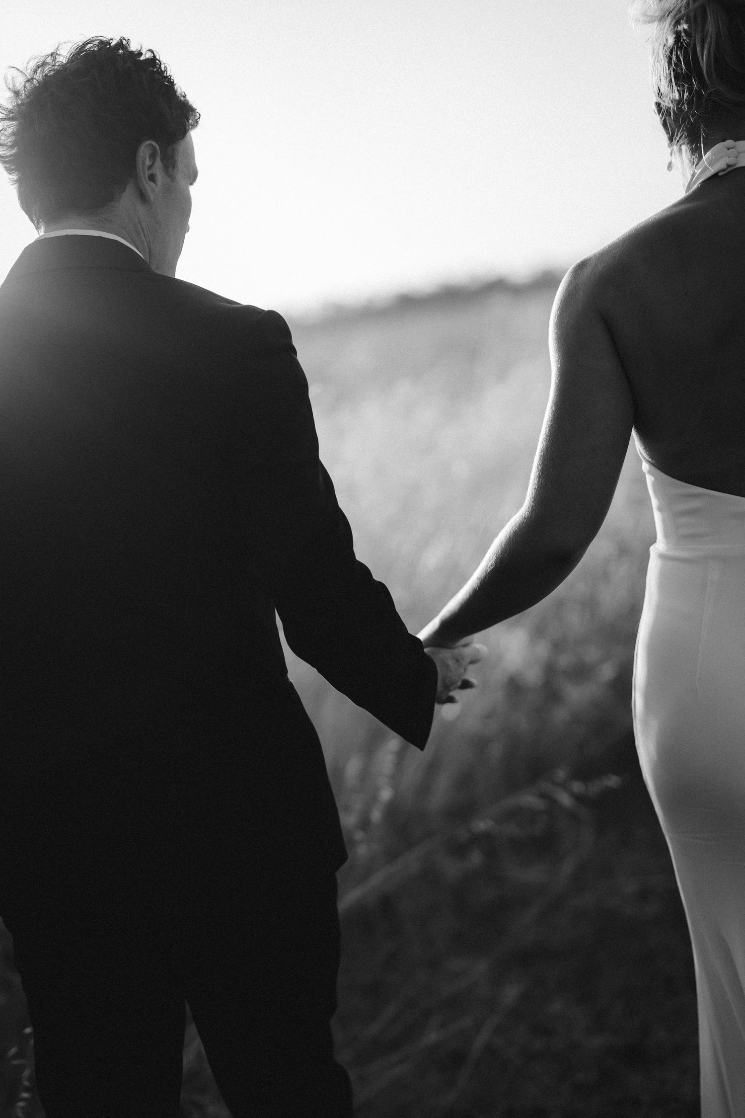 A black and white close-up of a couple holding hands while walking through tall grass.
