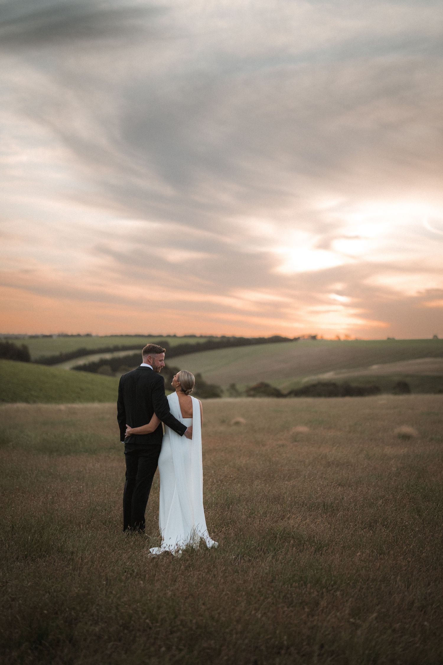 A couple in formal wedding attire embraces in a golden field at sunset.