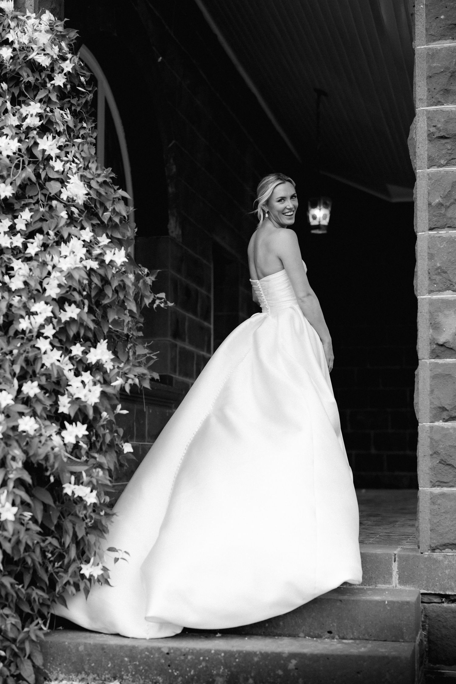 Bride in strapless ballgown wedding dress standing on stone steps, smiling over shoulder near flowering vines, black and white photo.