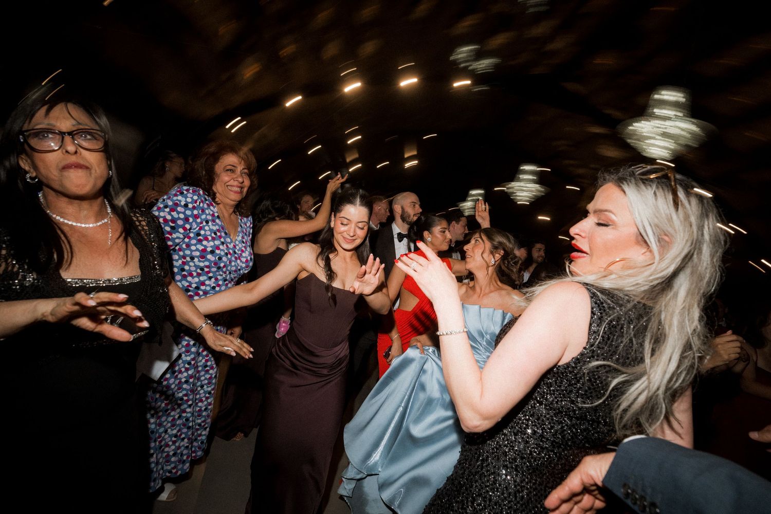 Wedding guests dance and celebrate joyfully on a crowded dance floor under warm string lights at an evening reception.