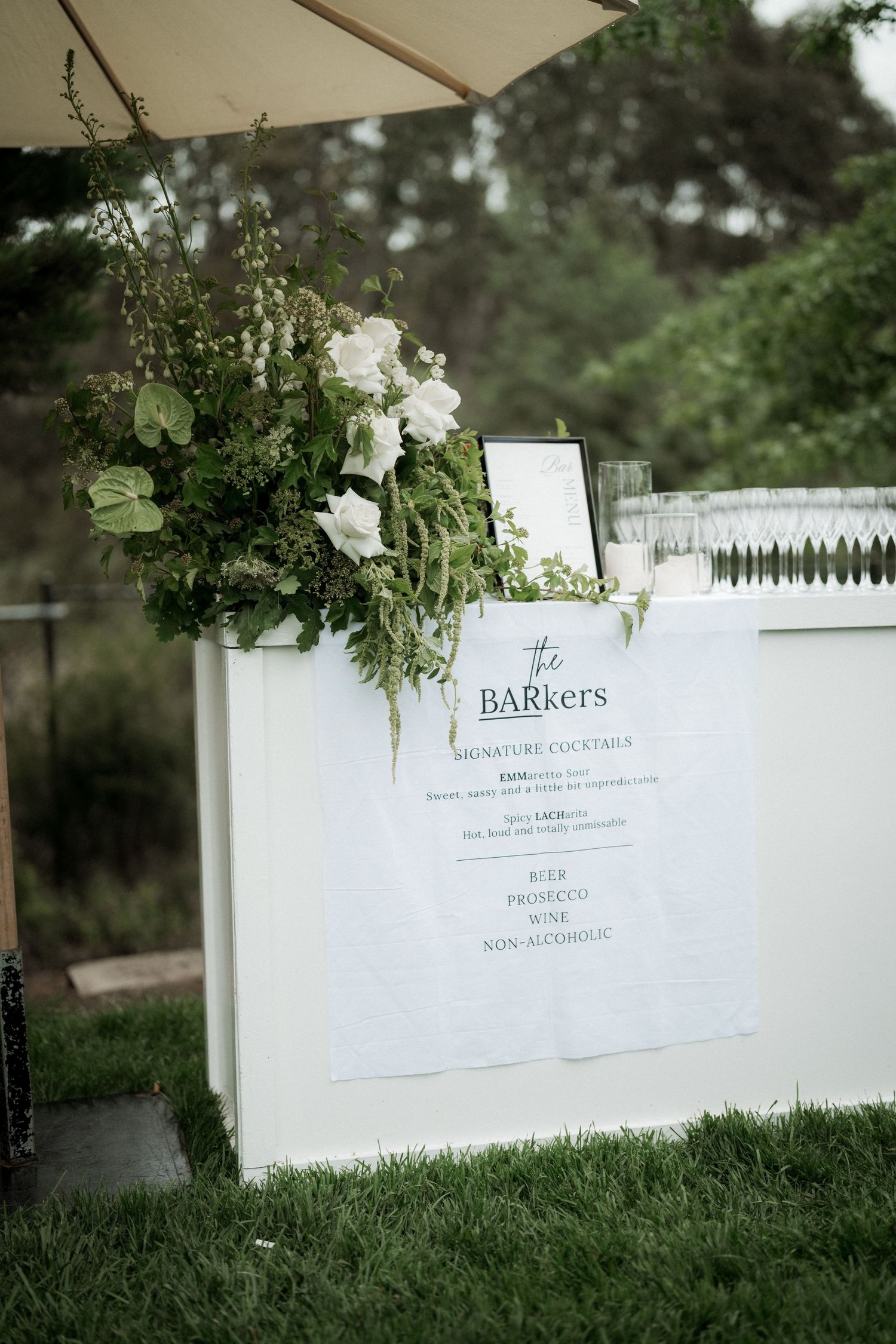 Elegant outdoor wedding bar with white floral arrangements, greenery, and a signature cocktail menu sign on grass.