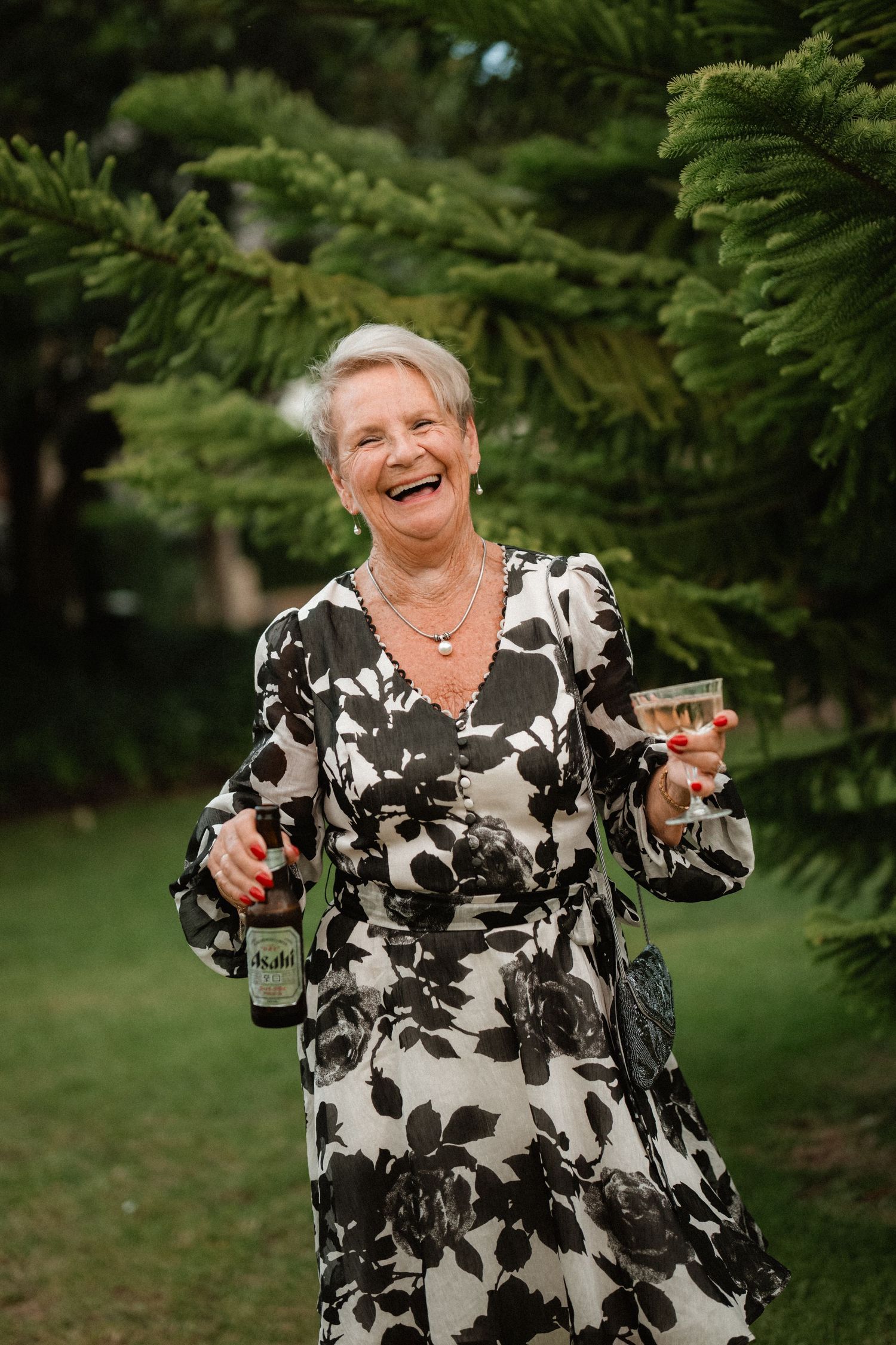 A guest in a floral dress laughs joyfully while holding wine at an outdoor wedding celebration.