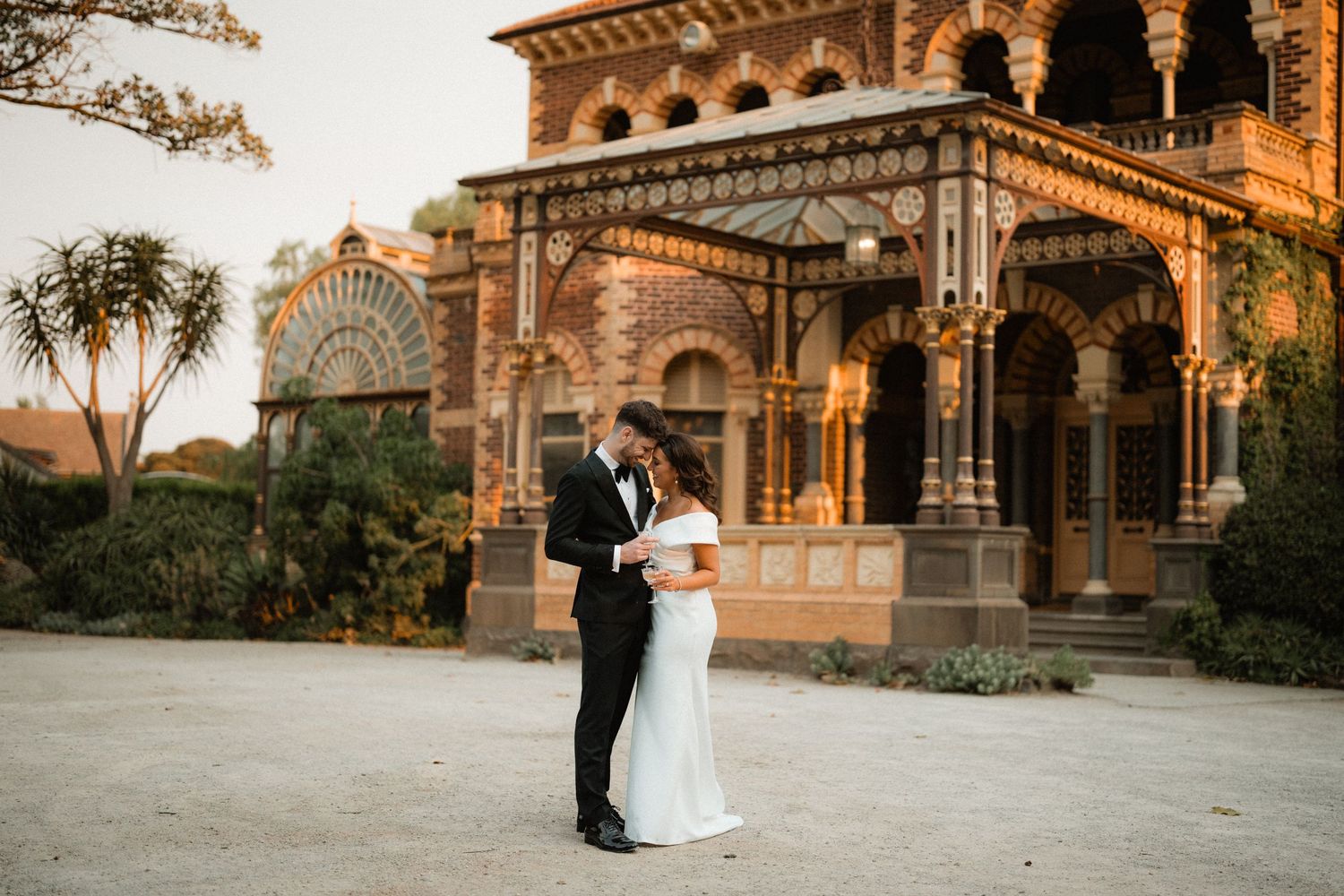 A couple celebrates their wedding at a majestic Victorian-style mansion with ornate architectural details at sunset.
