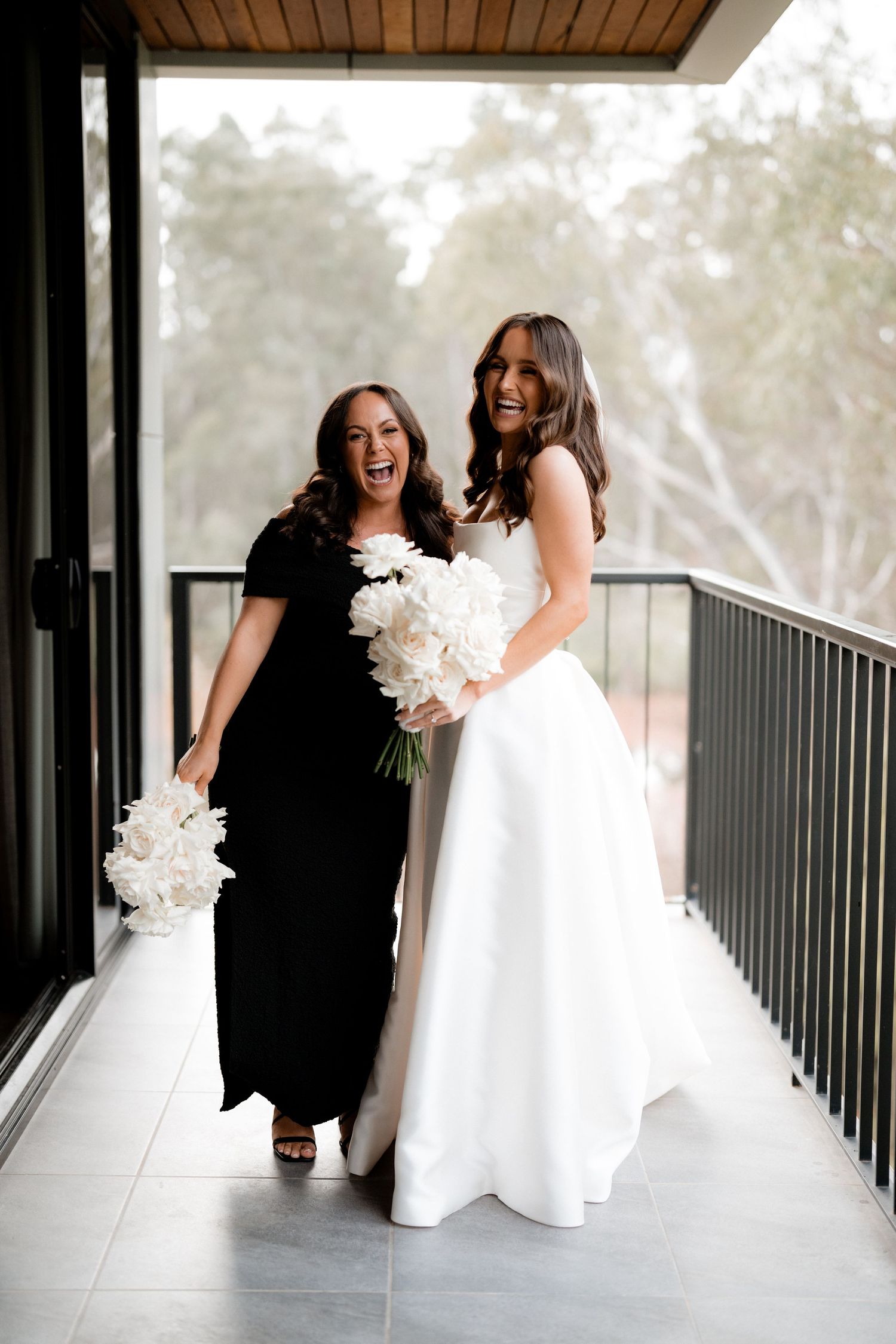 A bride and bridesmaid share a happy moment on a balcony with white floral bouquets.