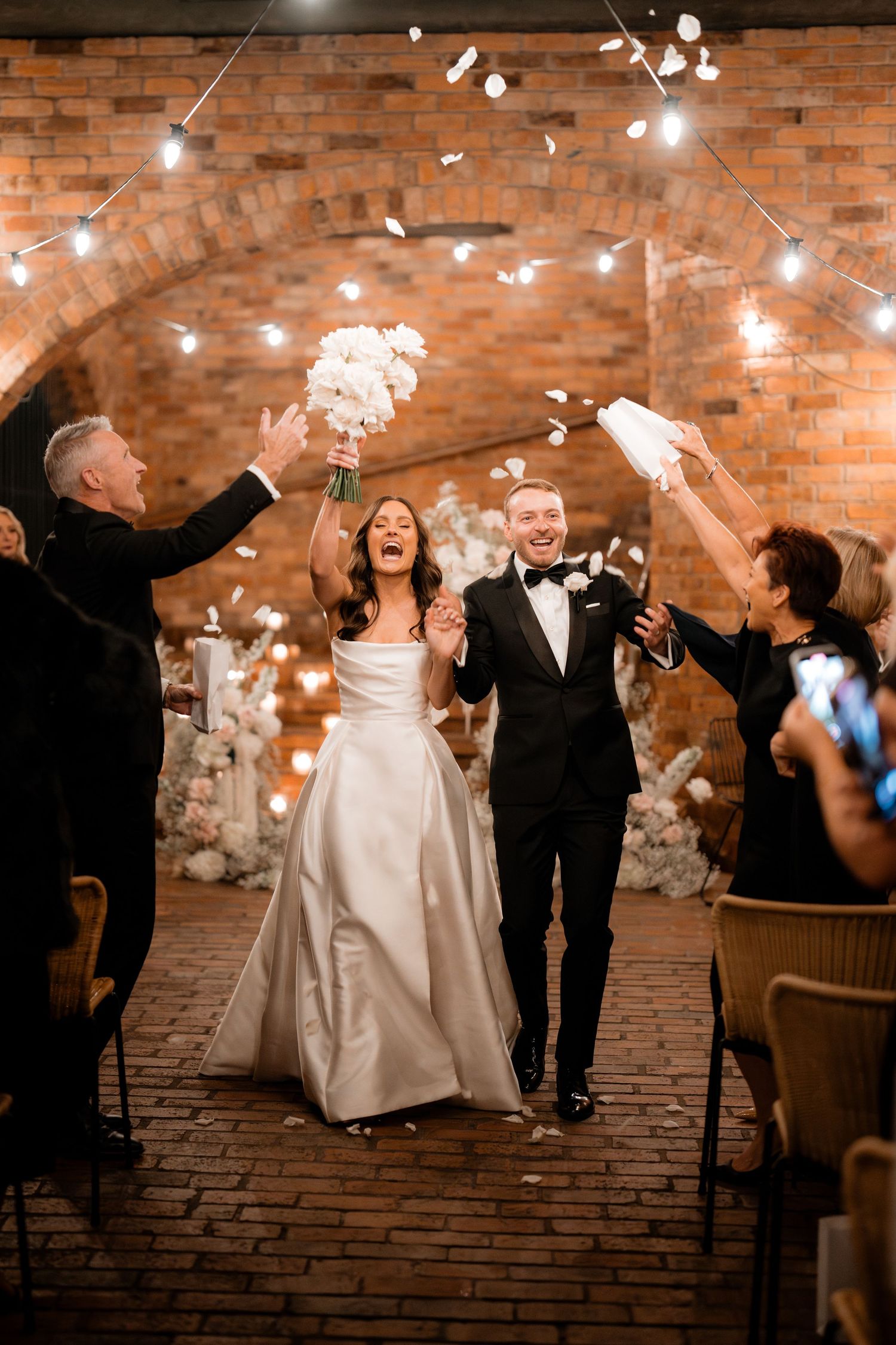 Wedding guests throw flower petals at smiling couple under brick archway with string lights.