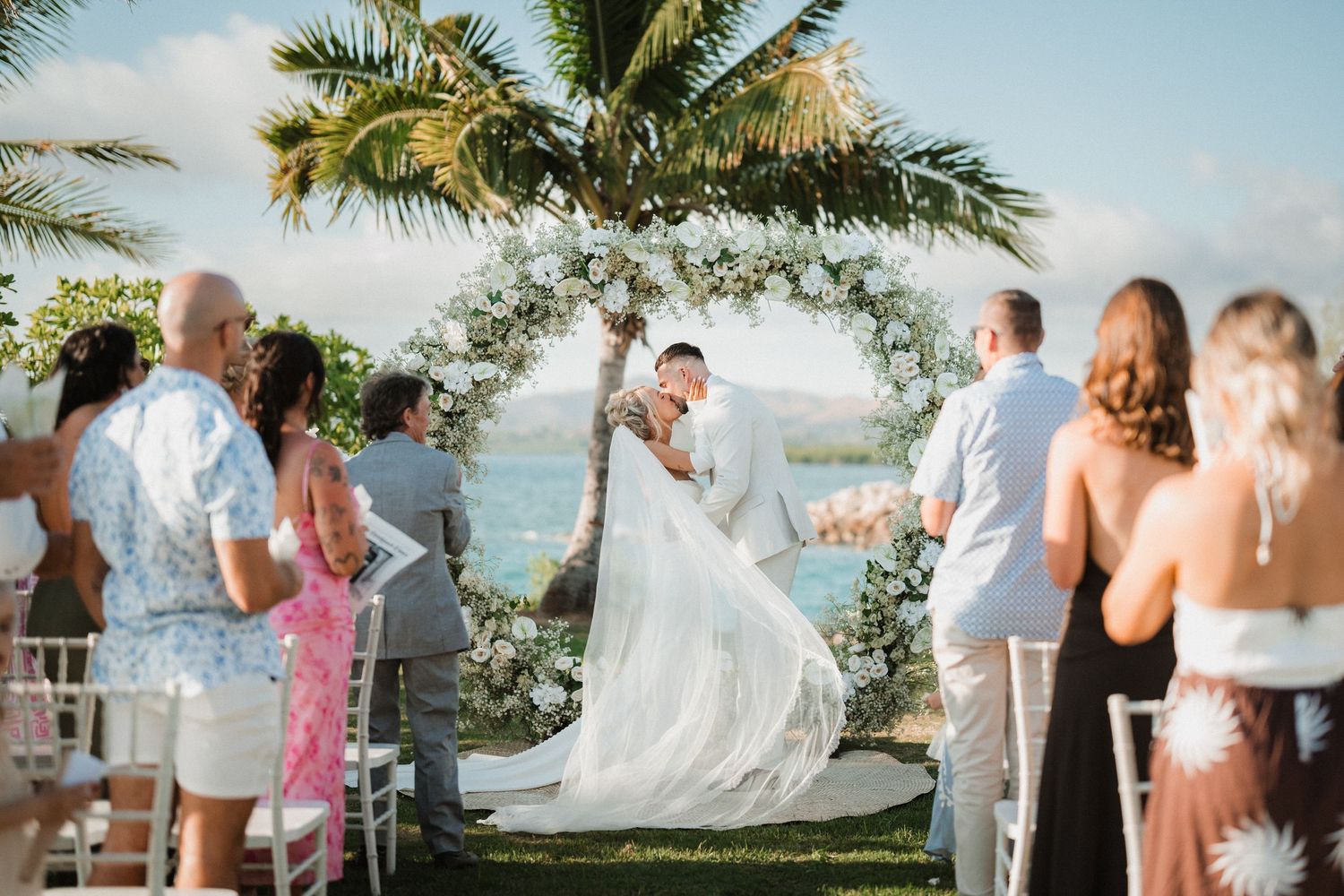 Tropical beach wedding ceremony under a floral arch with palm trees and ocean views.