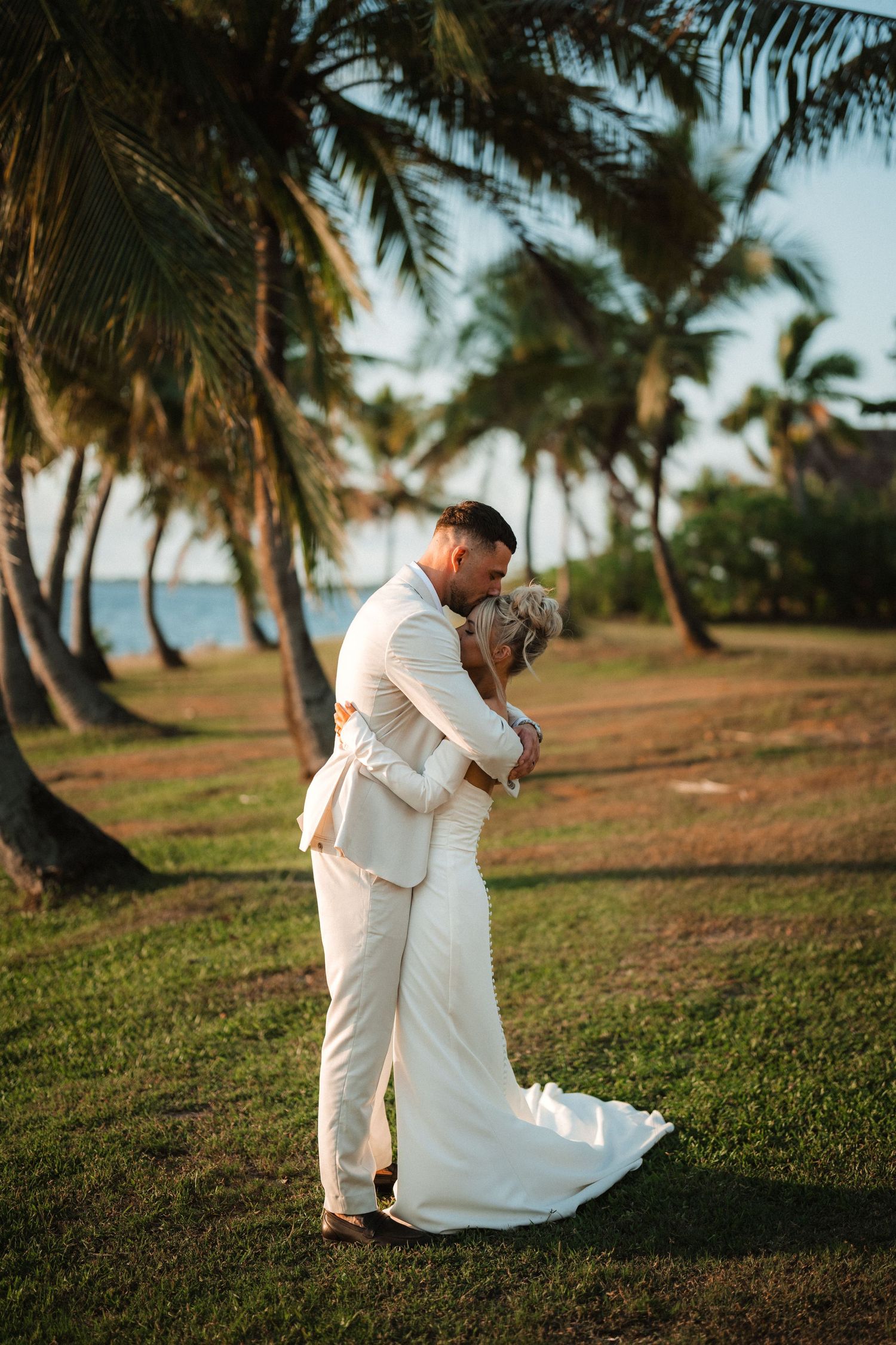 A romantic wedding couple embracing on a tropical beach lined with palm trees at sunset.