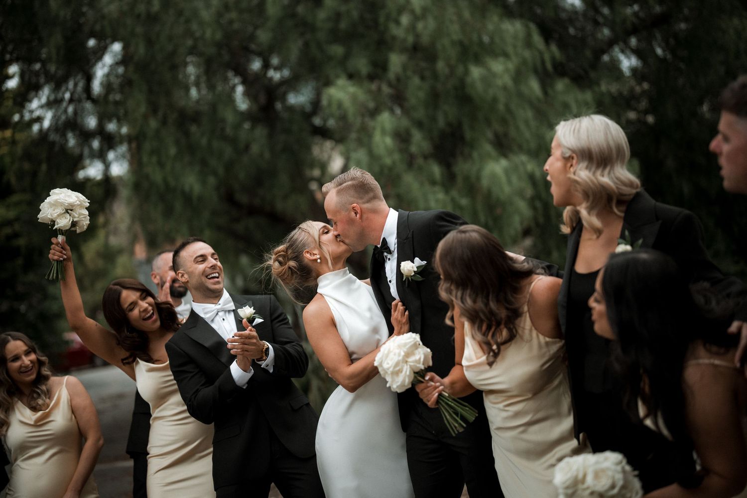 Wedding party dressed in black and ivory formal attire laughing joyfully together outdoors under trees.