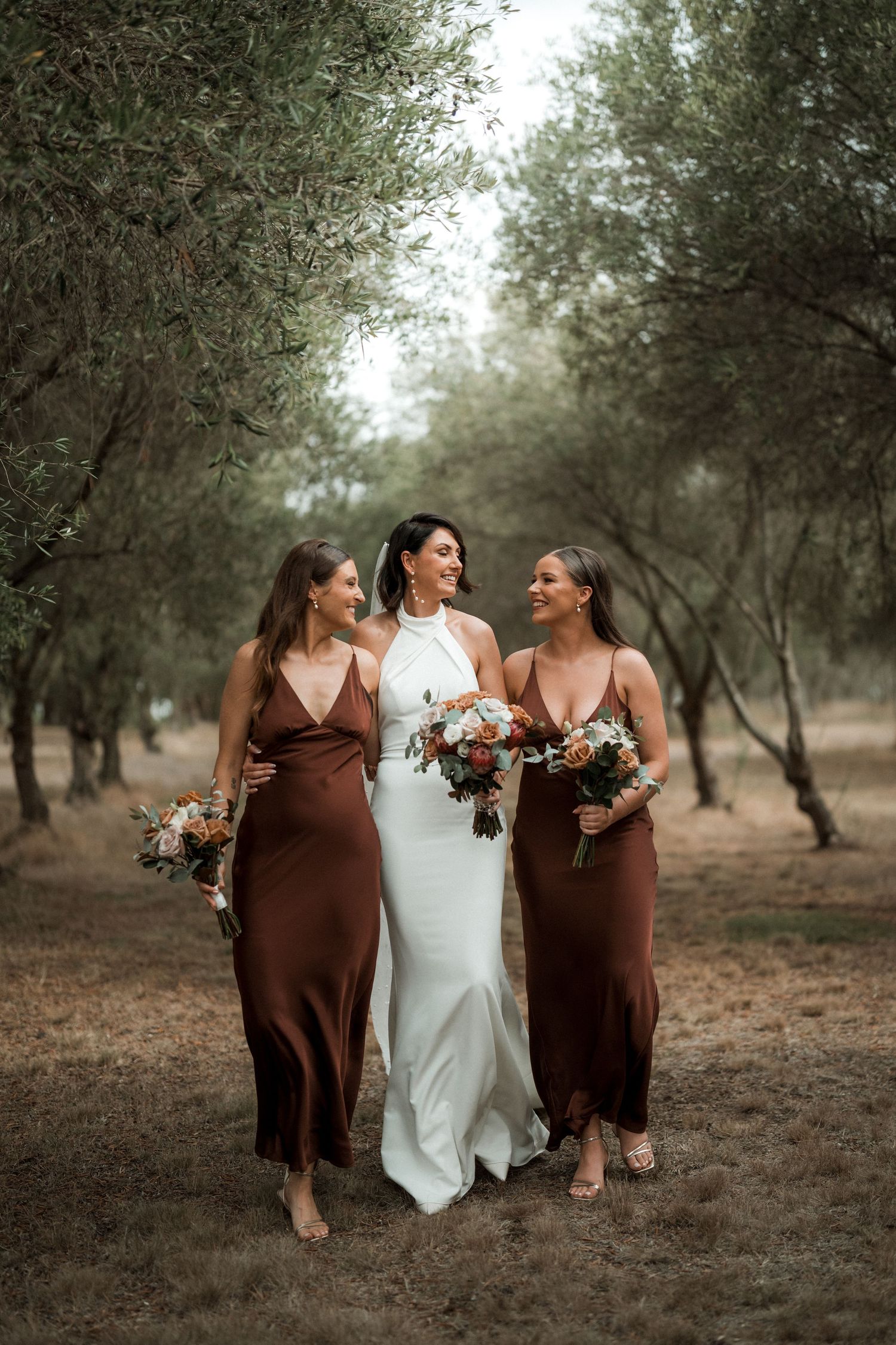 Three women in white and brown dresses holding bouquets walk together through an olive grove.