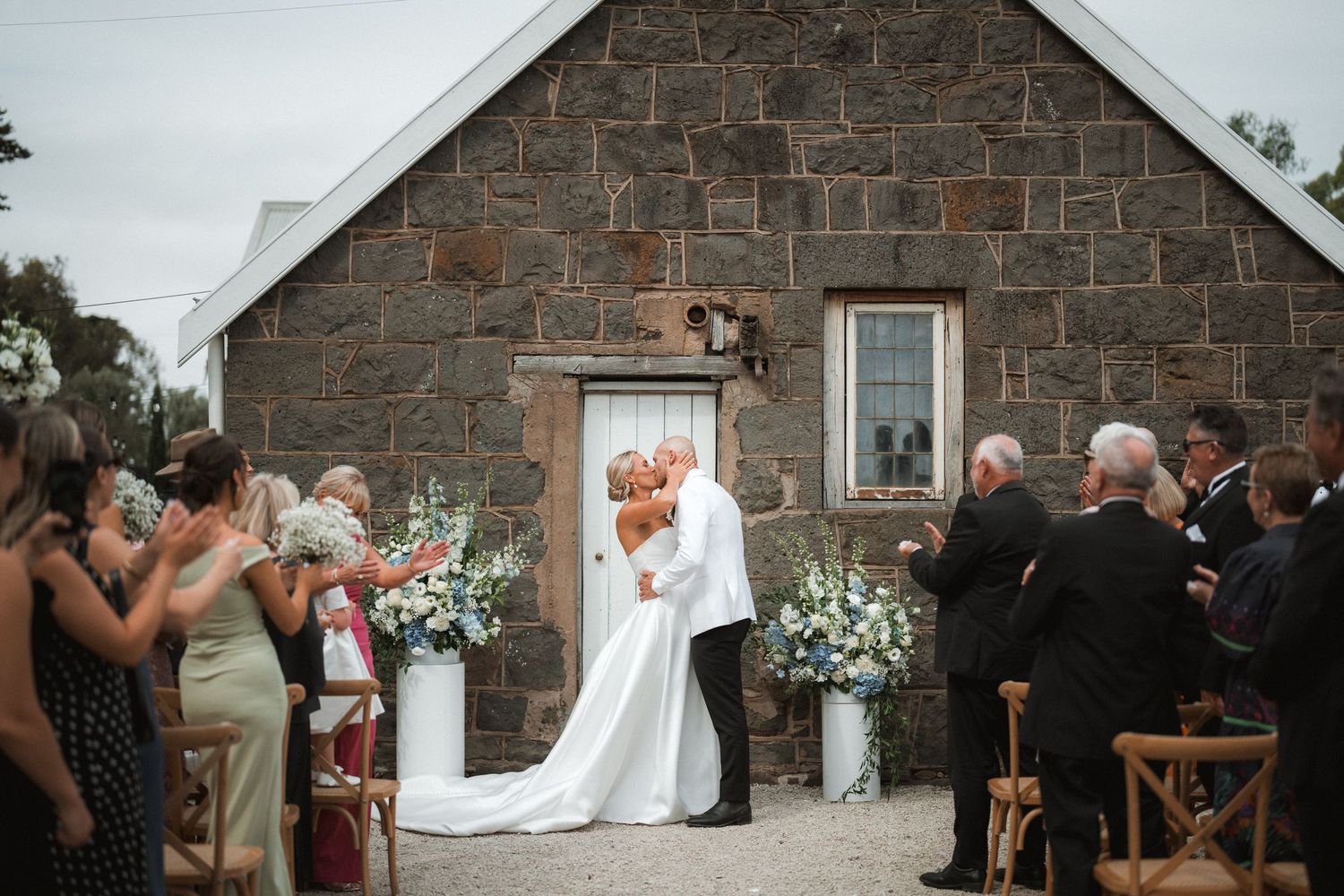 A stone farmhouse wedding ceremony with floral arrangements and wooden chairs on a rustic outdoor setting.