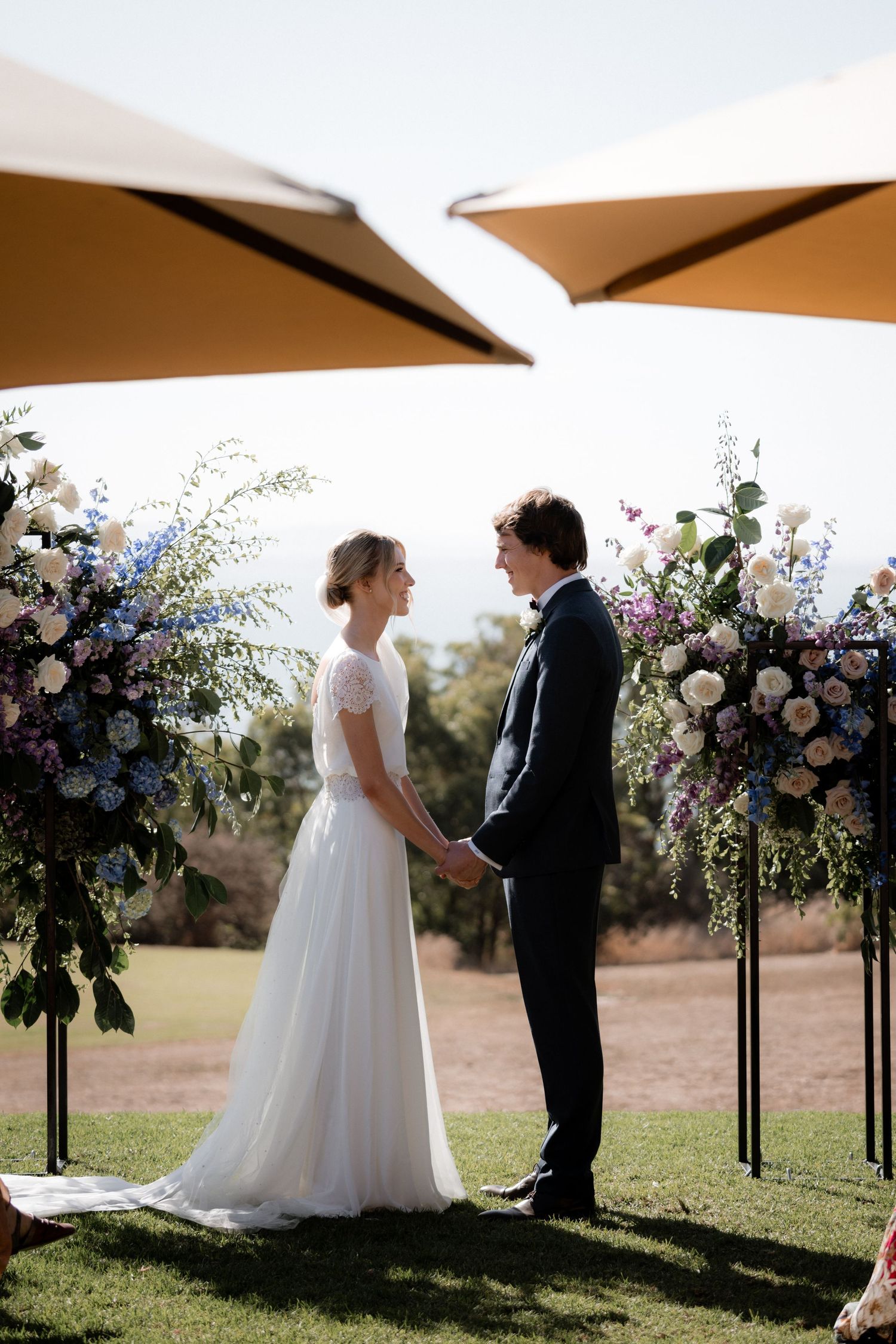 Wedding ceremony setup outdoors with elegant floral arrangements and beige umbrellas providing shade on a sunny day.