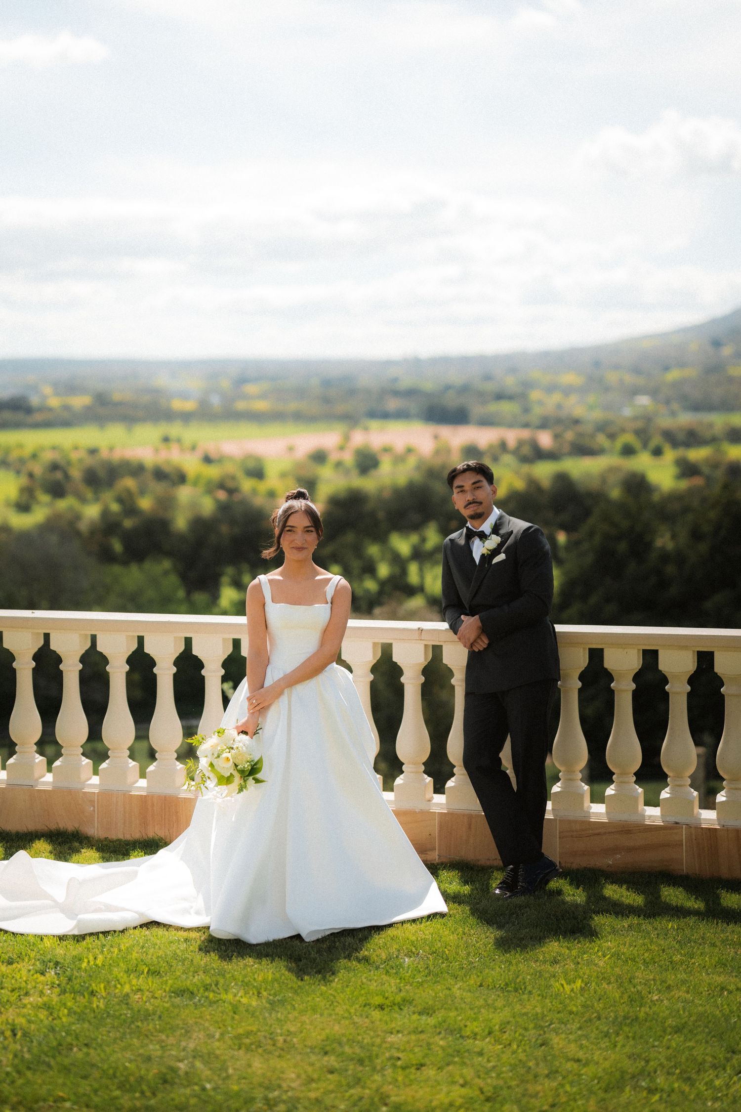 Bride in an elegant white gown holding a bouquet poses with her groom beside a stone balustrade overlooking green countryside.