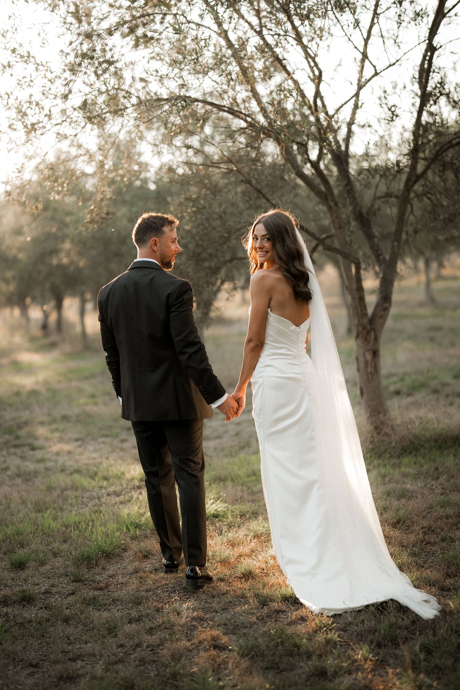 Romantic wedding photo of couple walking hand in hand through an olive grove at golden hour.