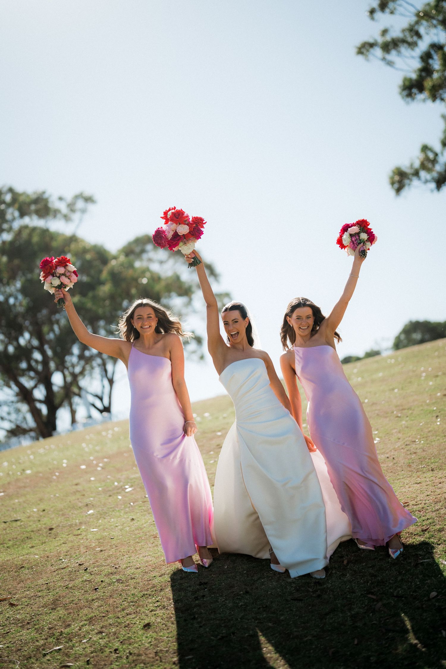 Bridesmaids in pink dresses and bride in white gown sit playfully on grassy hillside with bouquets.