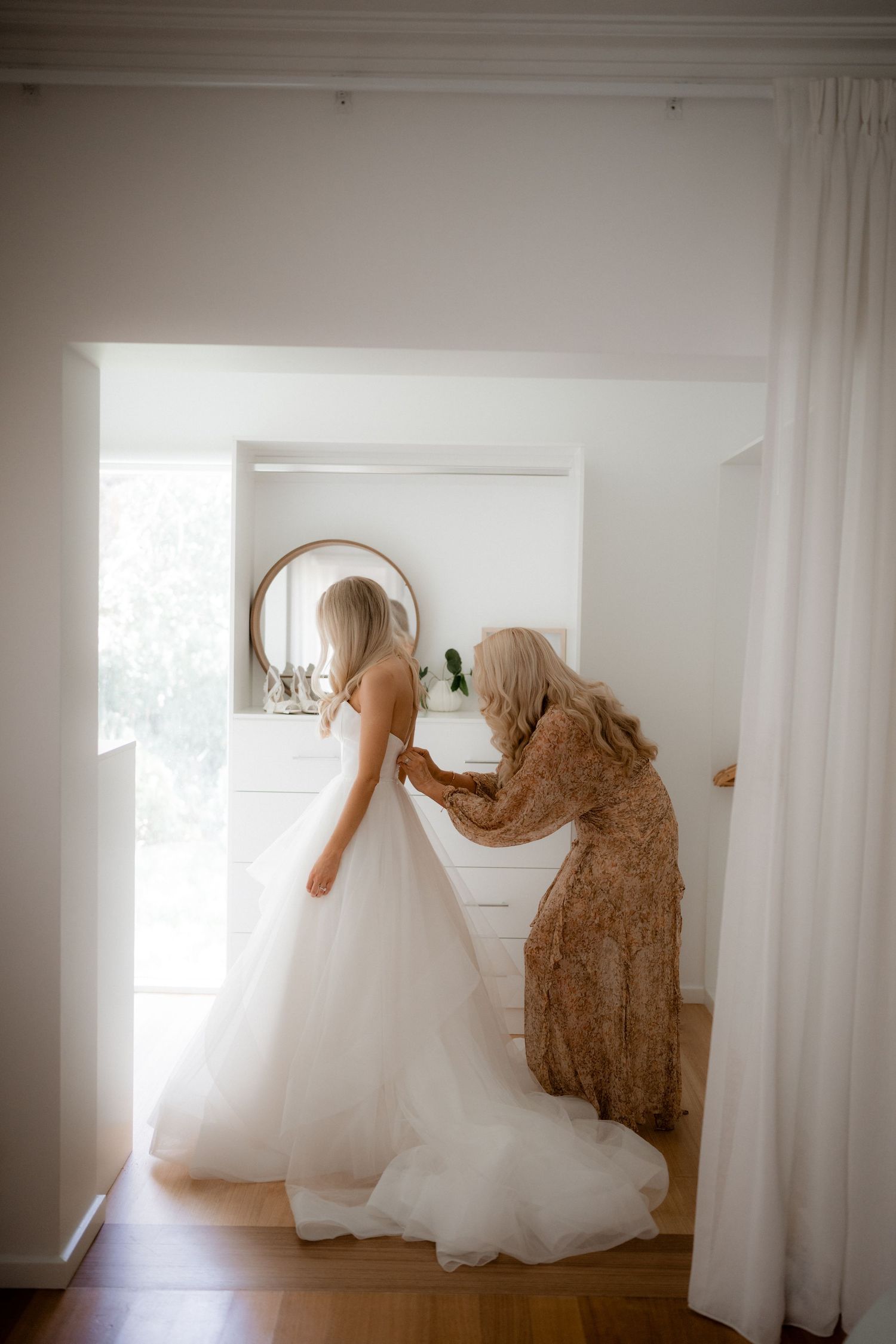 A flowing white wedding dress hangs elegantly in a bright sunlit room with wooden floors.