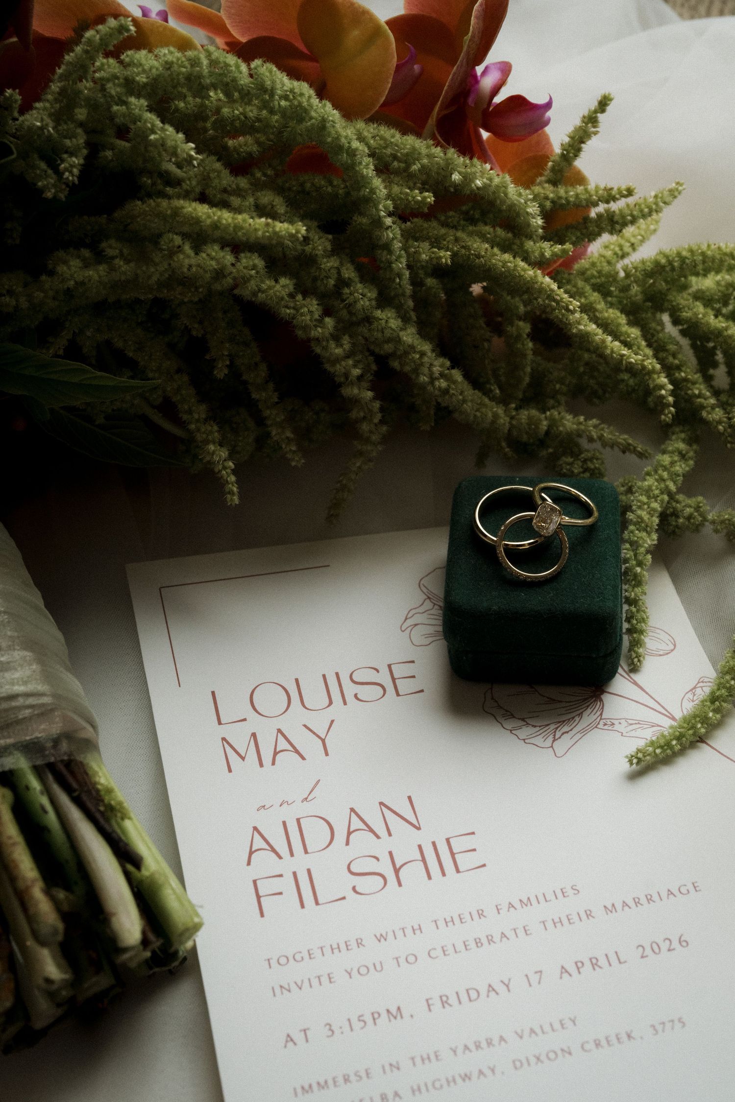 Wedding invitation for Louise May and Aidan Filshie with gold rings on green velvet box and floral bouquet.